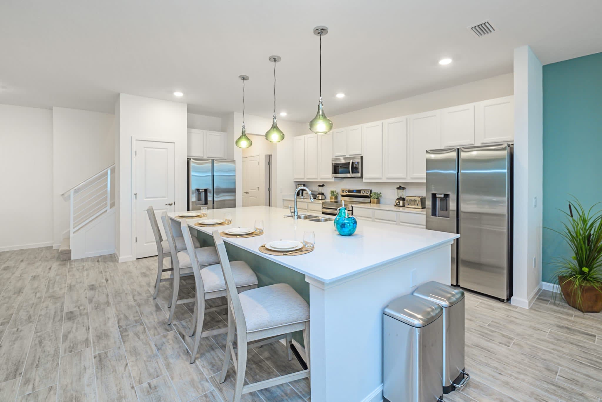 Modern kitchen featuring a large white island with a double sink and seating for four at bar stools. White shaker cabinets, stainless steel appliances, and light green pendant lights complete the bright and functional space.