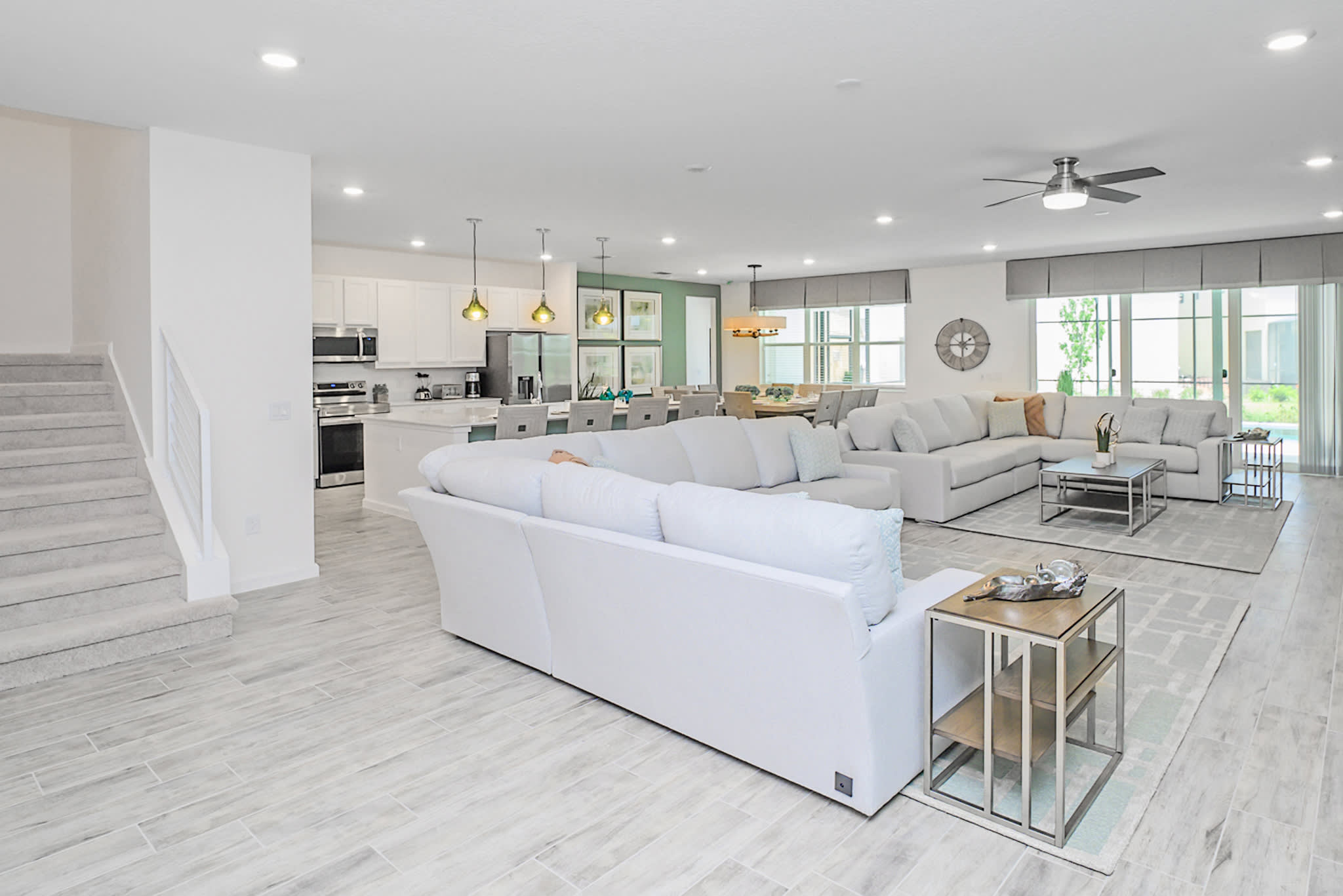 Expansive open-concept living space showcasing a large white sectional sofa in the foreground, leading to the dining area and a modern white kitchen. Light wood-look tile flooring unifies the entire area, with a staircase visible on the left.