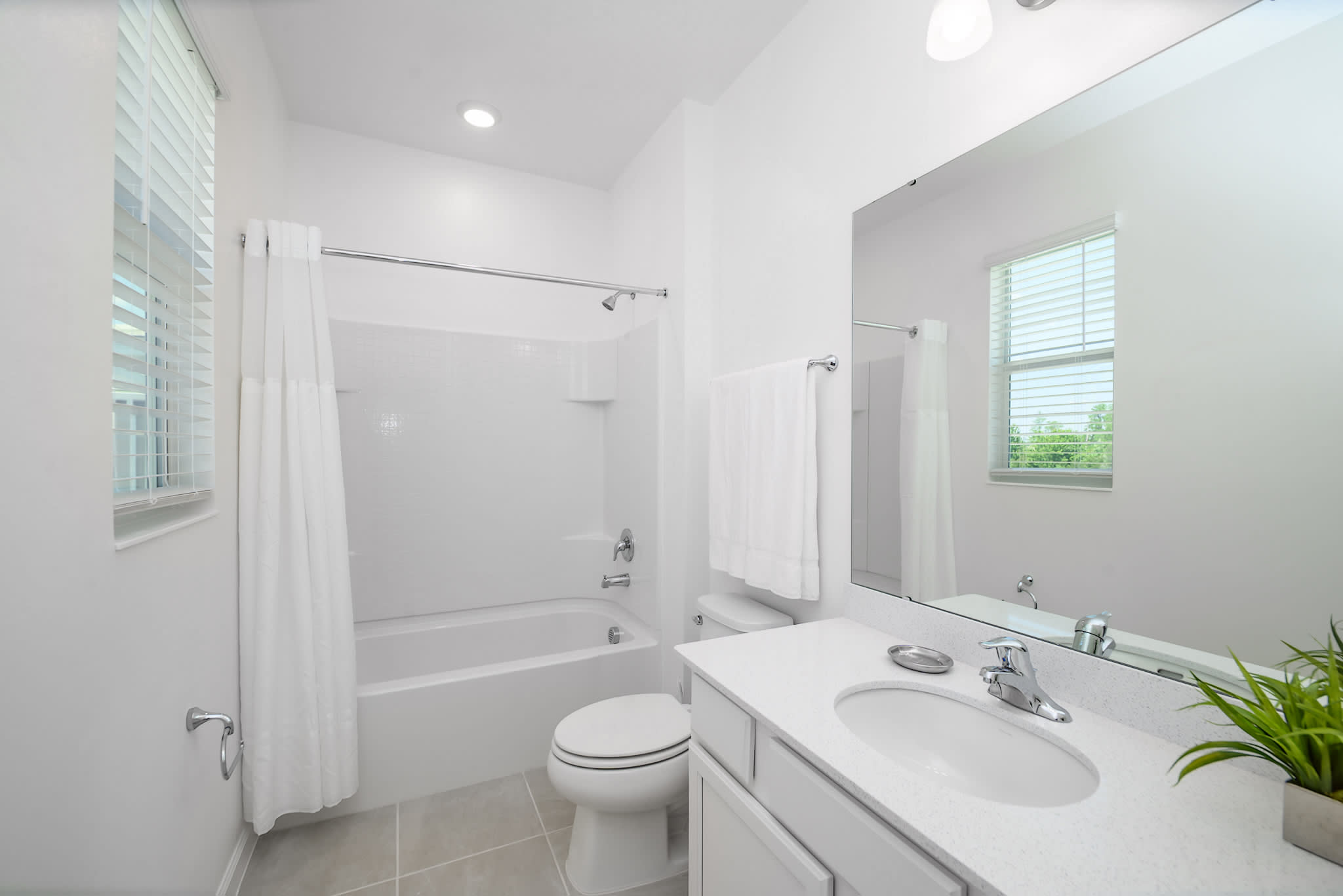 A bright bathroom with a window featuring blinds, a bathtub with a shower curtain, and a white toilet. A vanity with a white countertop and sink is on the right, adorned with a small plant.