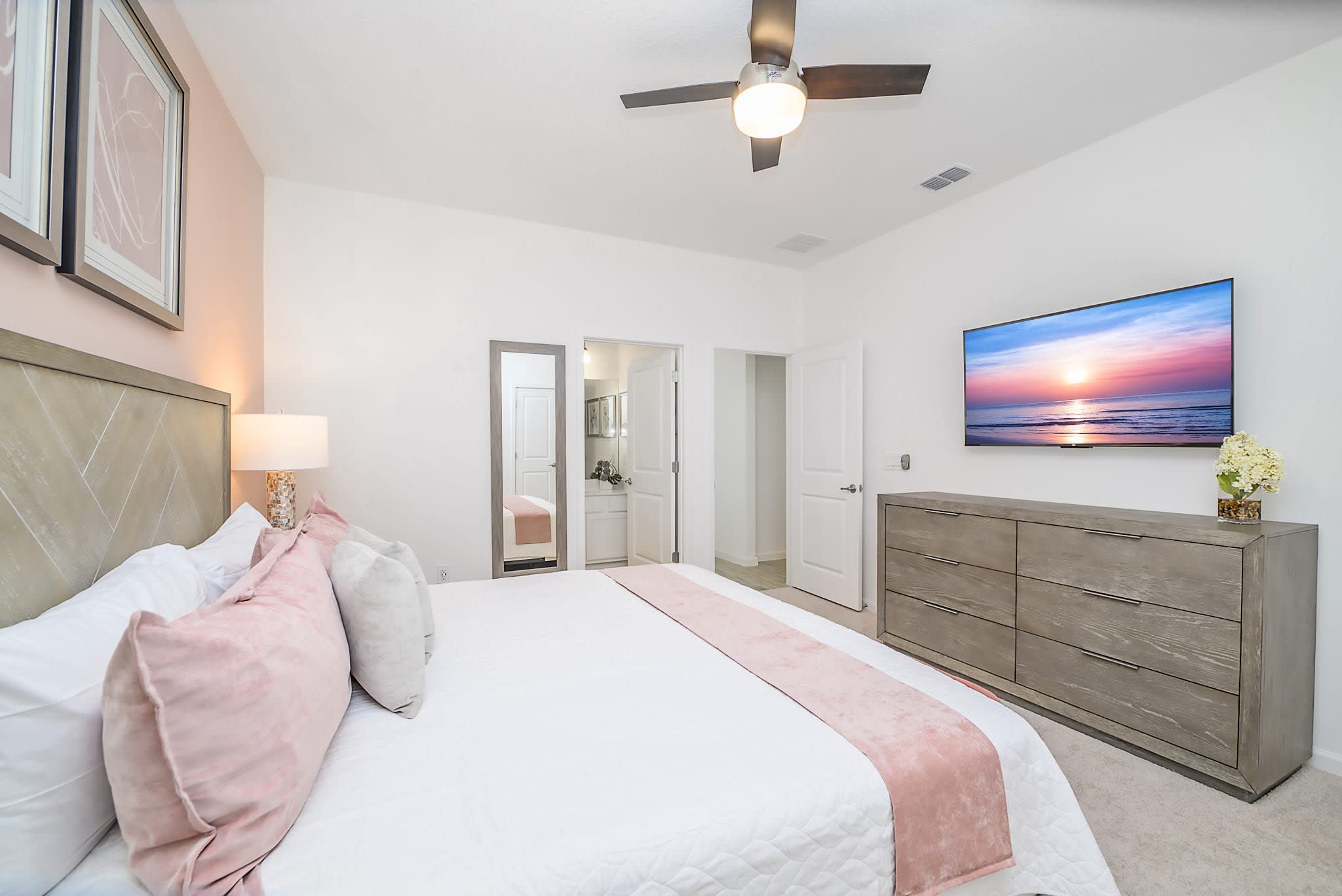 A stylish bedroom showcasing a queen-sized bed with pink decorative pillows and a matching throw, a gray wooden dresser with a wall-mounted TV, and a full-length mirror near the bathroom entrance.