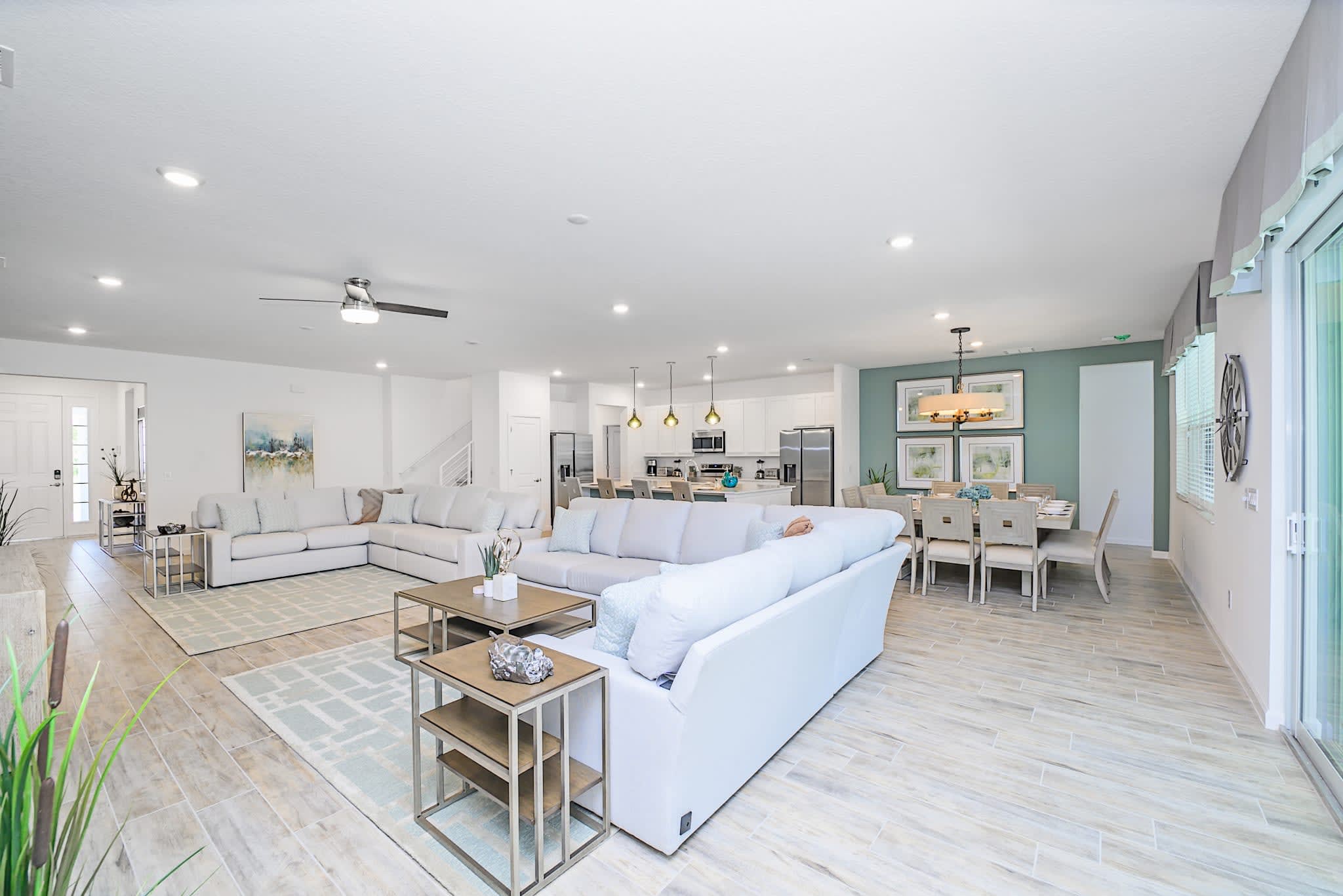Spacious open-concept living area featuring two large white sectional sofas arranged around coffee tables on a light-colored rug. Light wood-look tile flooring extends throughout, with a dining area and kitchen visible in the background.