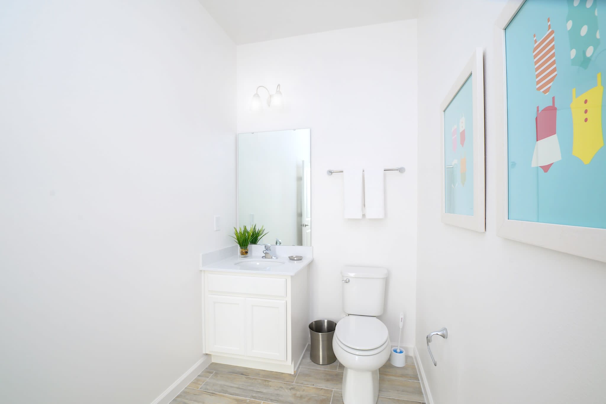 Clean white half bathroom with light wood-look tile flooring, a single vanity with a mirror, a toilet, and colorful framed artwork depicting swimsuits. The room is minimalist and bright.