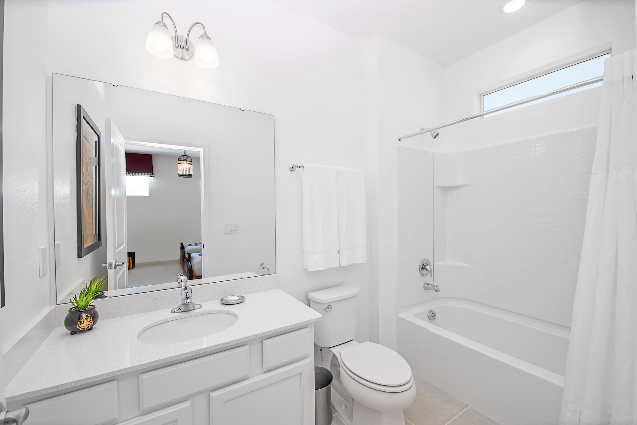 A clean, white bathroom with a single vanity, large mirror, toilet, and a bathtub with a shower. The room features white tiled walls and a small horizontal window above the shower for natural light.