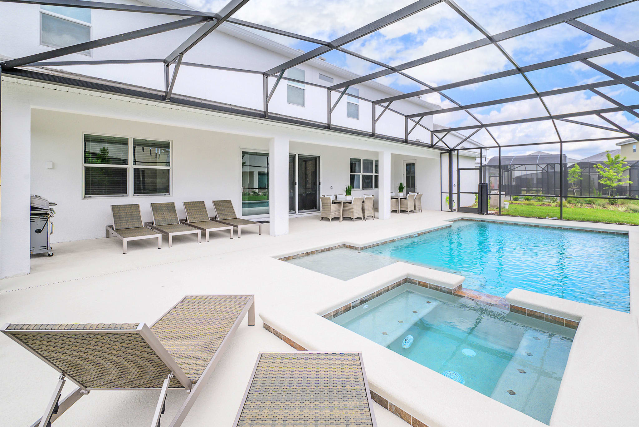 An expansive screened-in outdoor pool area featuring a large rectangular swimming pool and two integrated hot tubs. Several lounge chairs and an outdoor dining set are arranged on the white patio, surrounded by a black screen enclosure.
