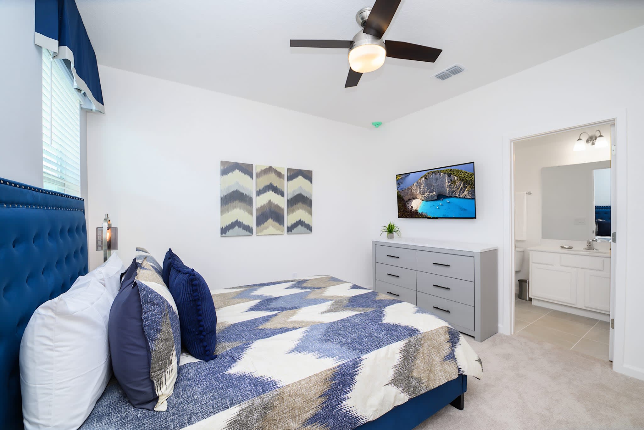 View of a modern bedroom with a wall-mounted TV above a gray dresser, facing a queen-sized bed with a blue upholstered headboard. A doorway leads to an en-suite bathroom, and decorative wall art is visible.