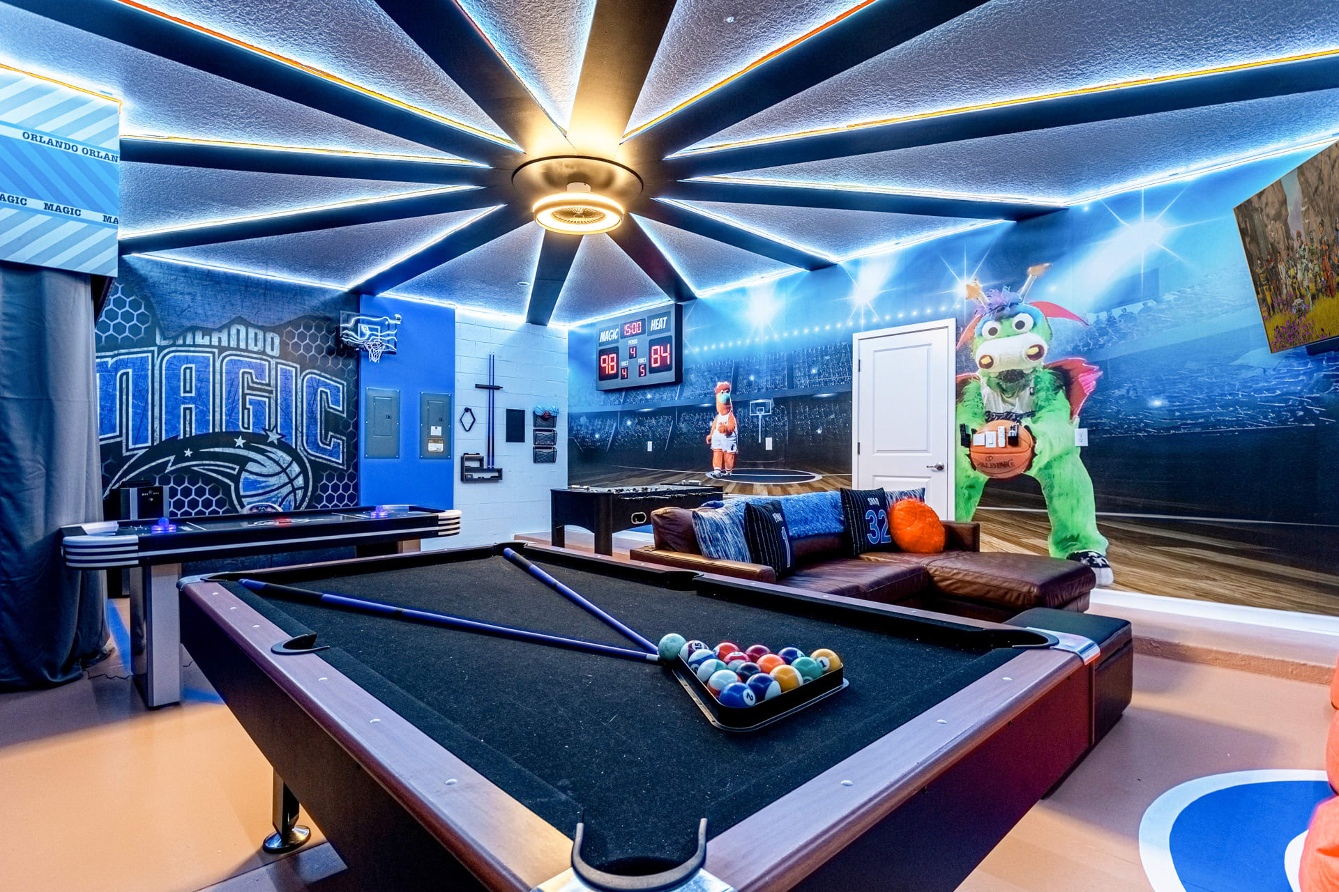 A vibrant game room featuring a pool table with colorful balls, a foosball table, and a basketball court-themed floor. The ceiling has radial LED lighting, and the walls are adorned with Orlando Magic branding and a stadium mural.