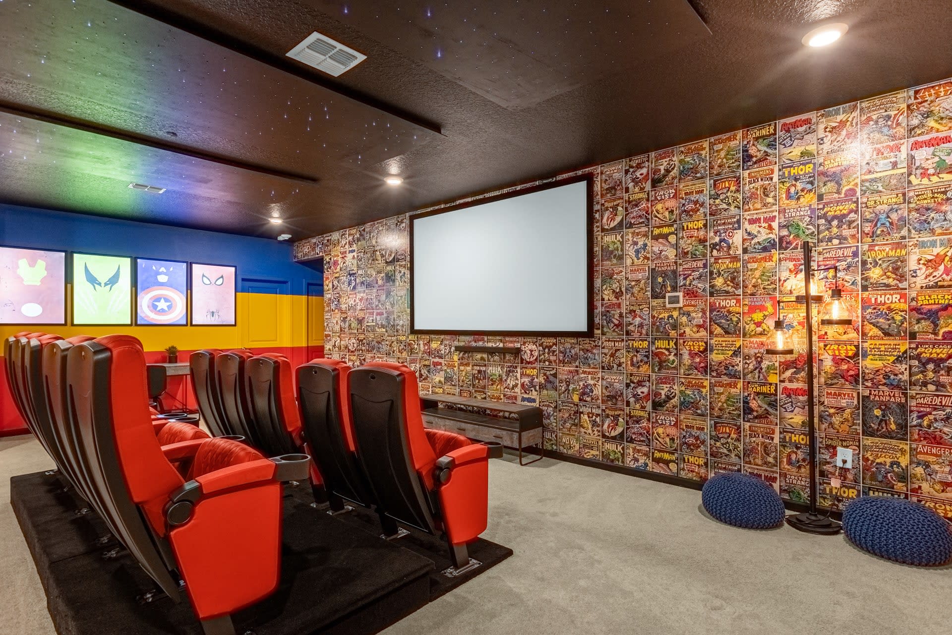 Another view of the media room, showcasing red tiered cinema seats and a large projection screen. The left wall is covered in colorful vintage comic book wallpaper, complementing the superhero theme.