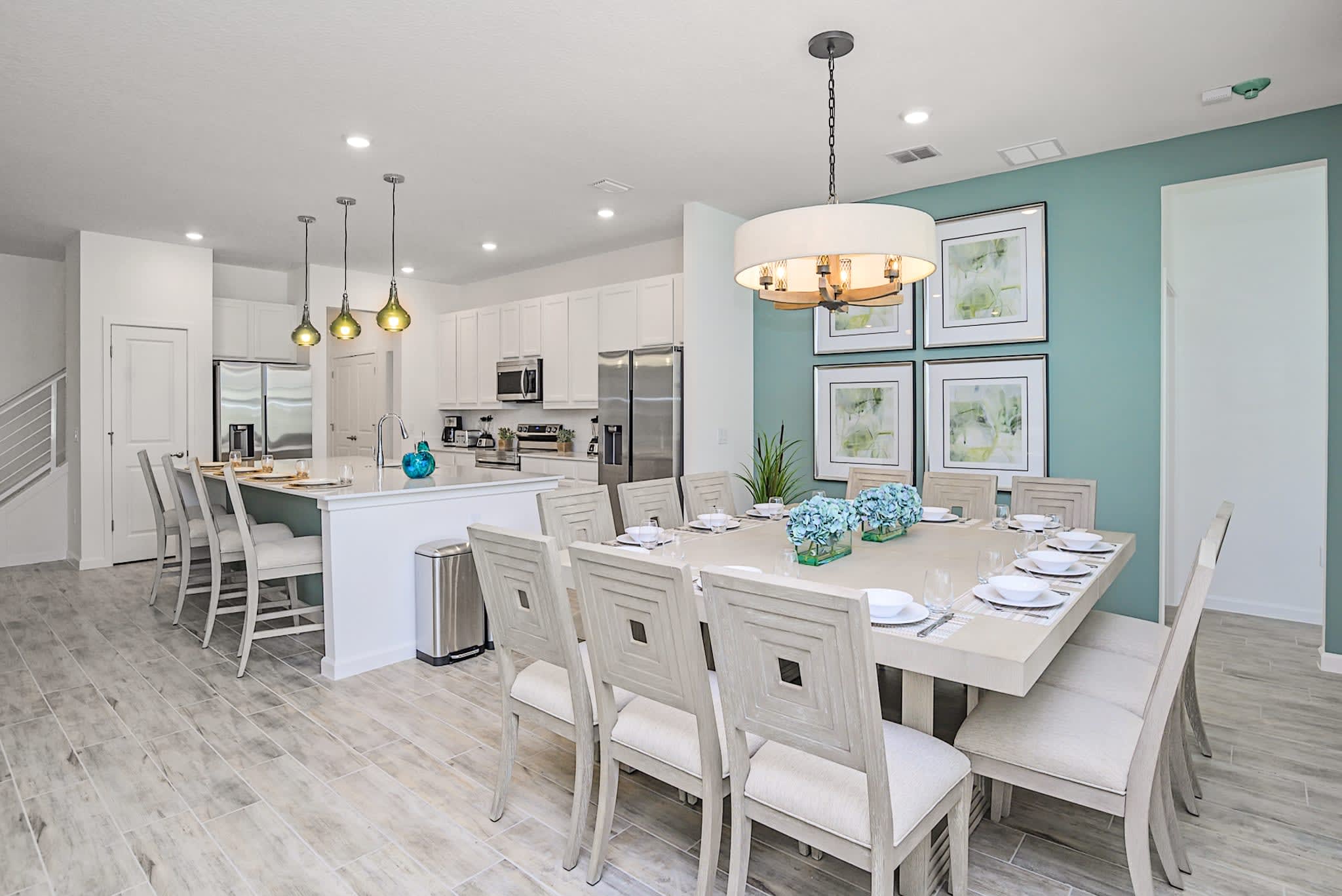 Elegant dining area featuring a large square dining table with seating for twelve, illuminated by a modern chandelier. A light teal accent wall with framed art adds a pop of color, with the bright white kitchen visible beyond.