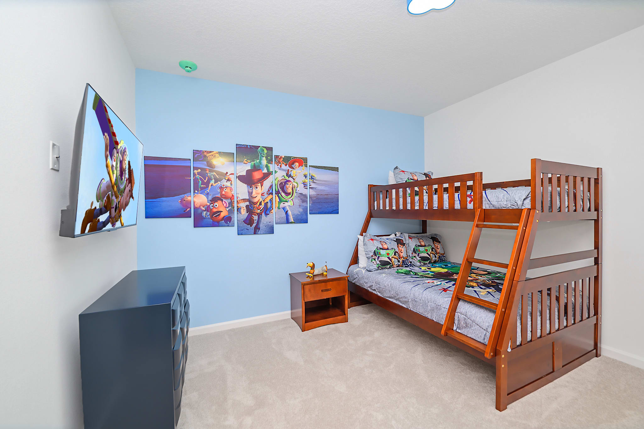 A Toy Story themed bedroom featuring a sturdy wooden bunk bed, a light blue accent wall, and large wall art depicting characters from Toy Story. The room also includes a dark dresser, a nightstand, and a wall-mounted TV.