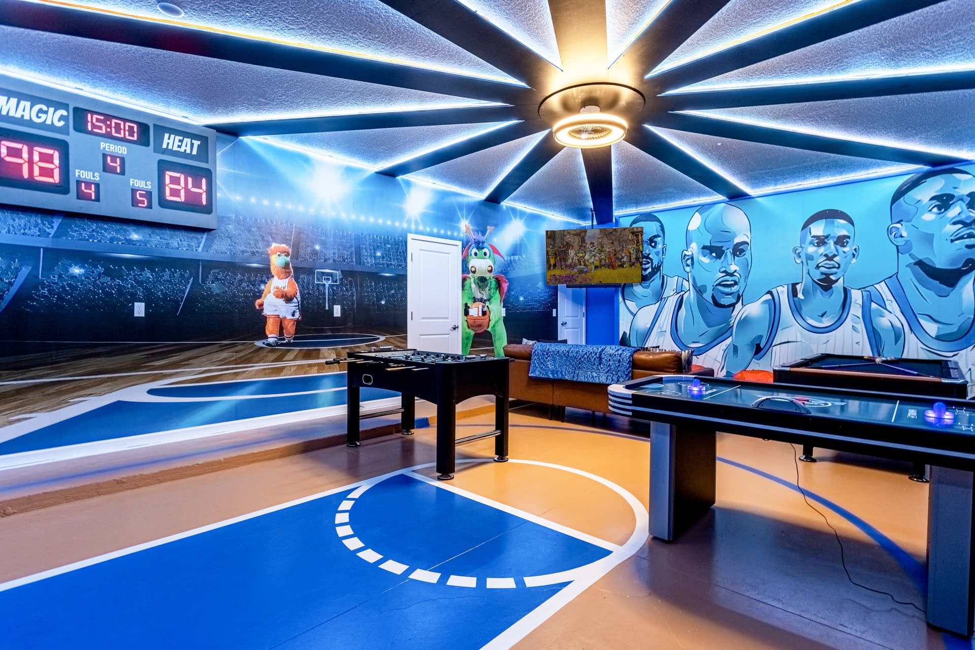 A dynamic game room with a basketball court-themed floor and a large mural of basketball players on the wall. The room includes a foosball table, an air hockey table, and a scoreboard displaying "Magic" vs "Heat" scores.