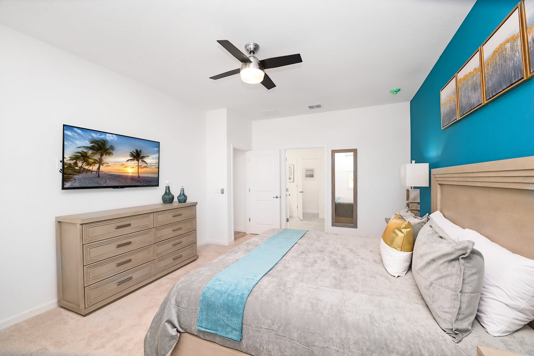 A modern bedroom featuring a king-sized bed with a light wood headboard, a vibrant blue accent wall, and a wall-mounted TV above a light wood dresser. A ceiling fan with a light fixture is centered in the room.
