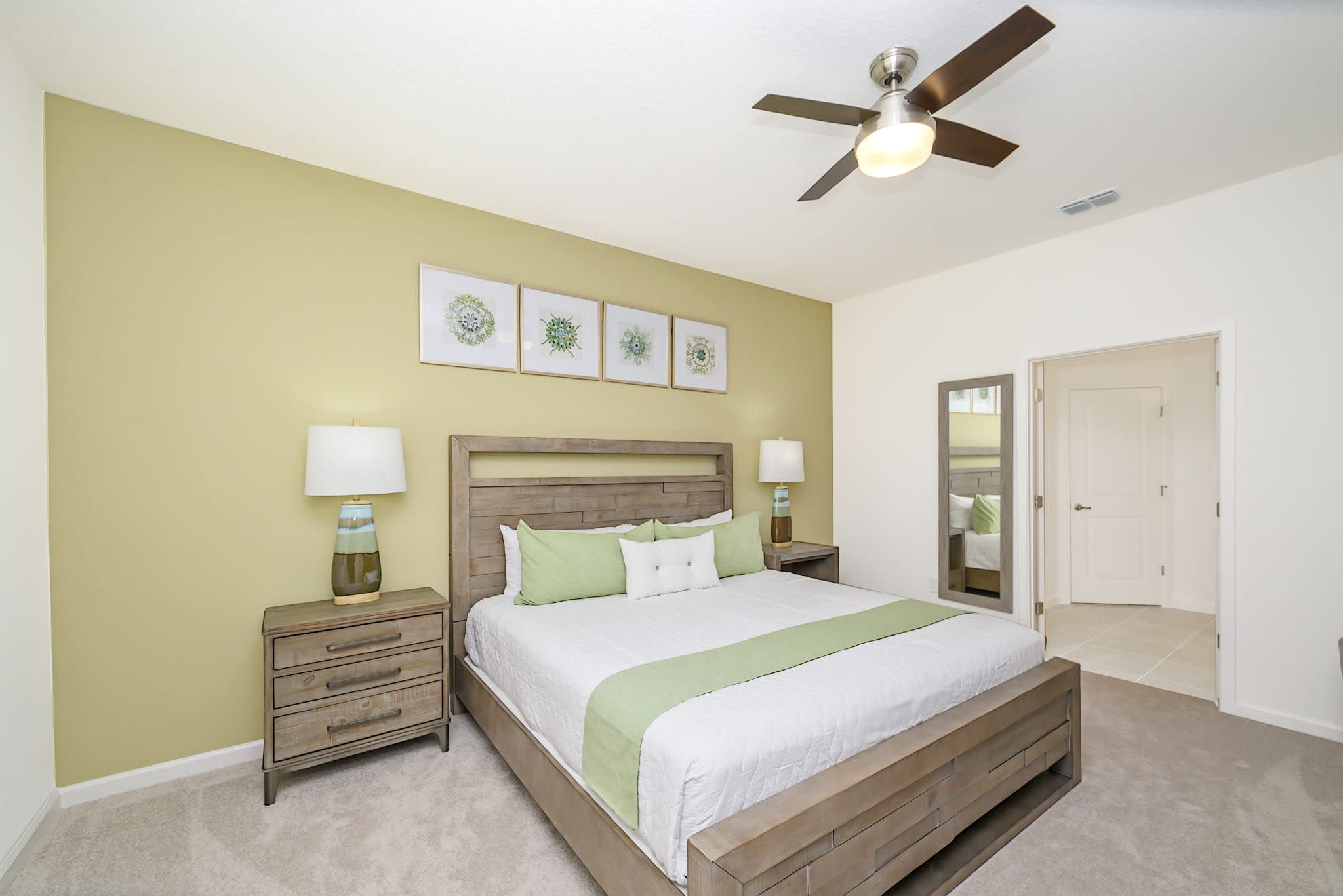 A cozy bedroom featuring a light wood king-sized bed with matching nightstands and lamps, set against a soft green accent wall. A full-length mirror reflects the room's entrance, enhancing the sense of space.