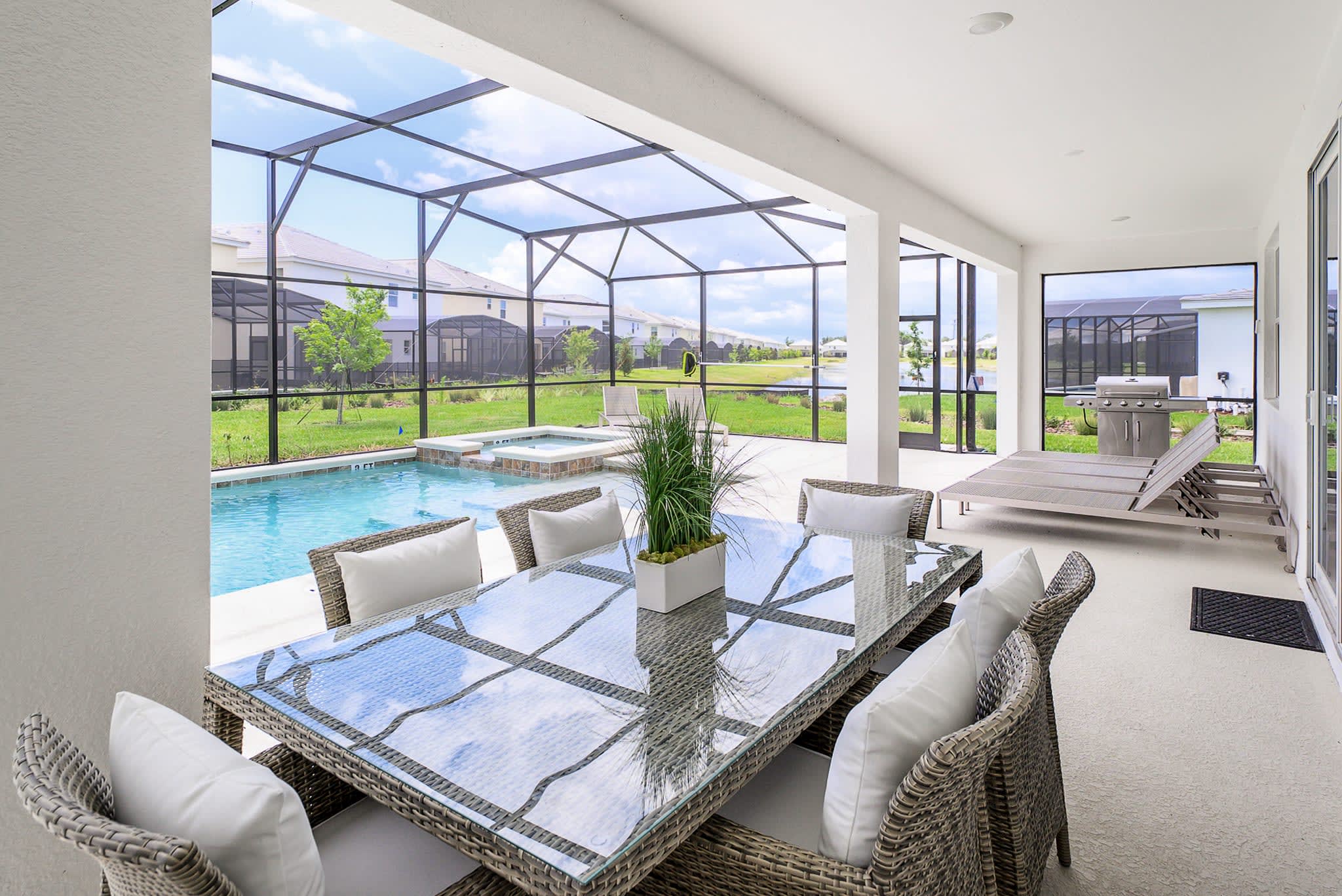 A covered outdoor patio area within the screen enclosure, featuring a large wicker dining table with a glass top and comfortable chairs. A BBQ grill is visible in the background, next to the swimming pool and hot tub.