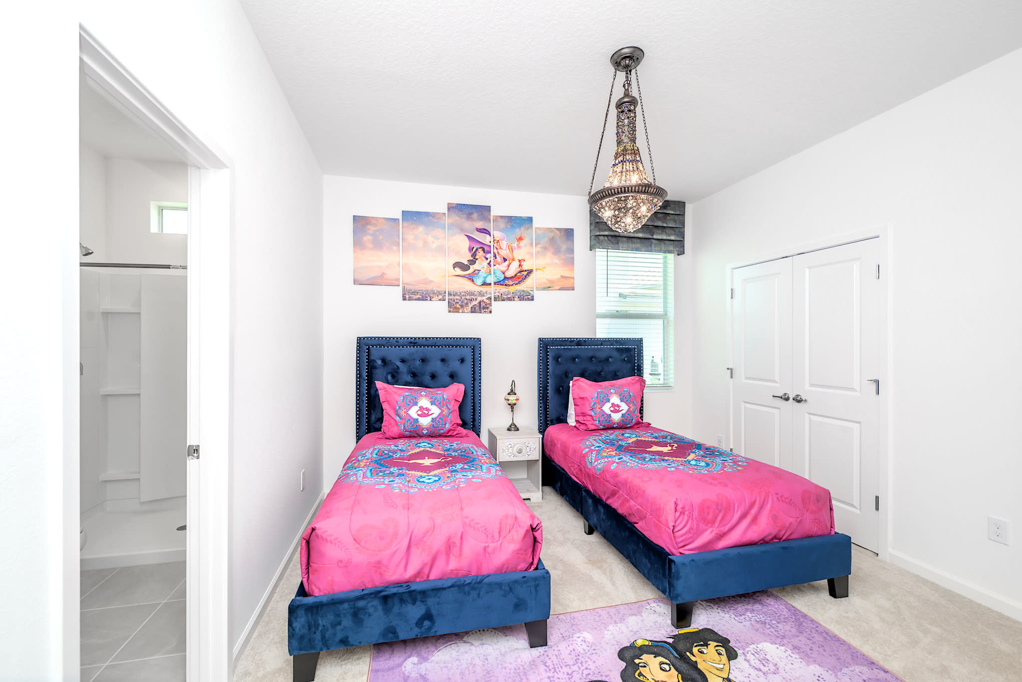 A vibrant themed bedroom with two navy blue velvet twin beds, each with bright pink Aladdin-themed bedding. A multi-panel wall art depicting Aladdin and Jasmine is above one bed, and an ornate chandelier hangs from the ceiling.
