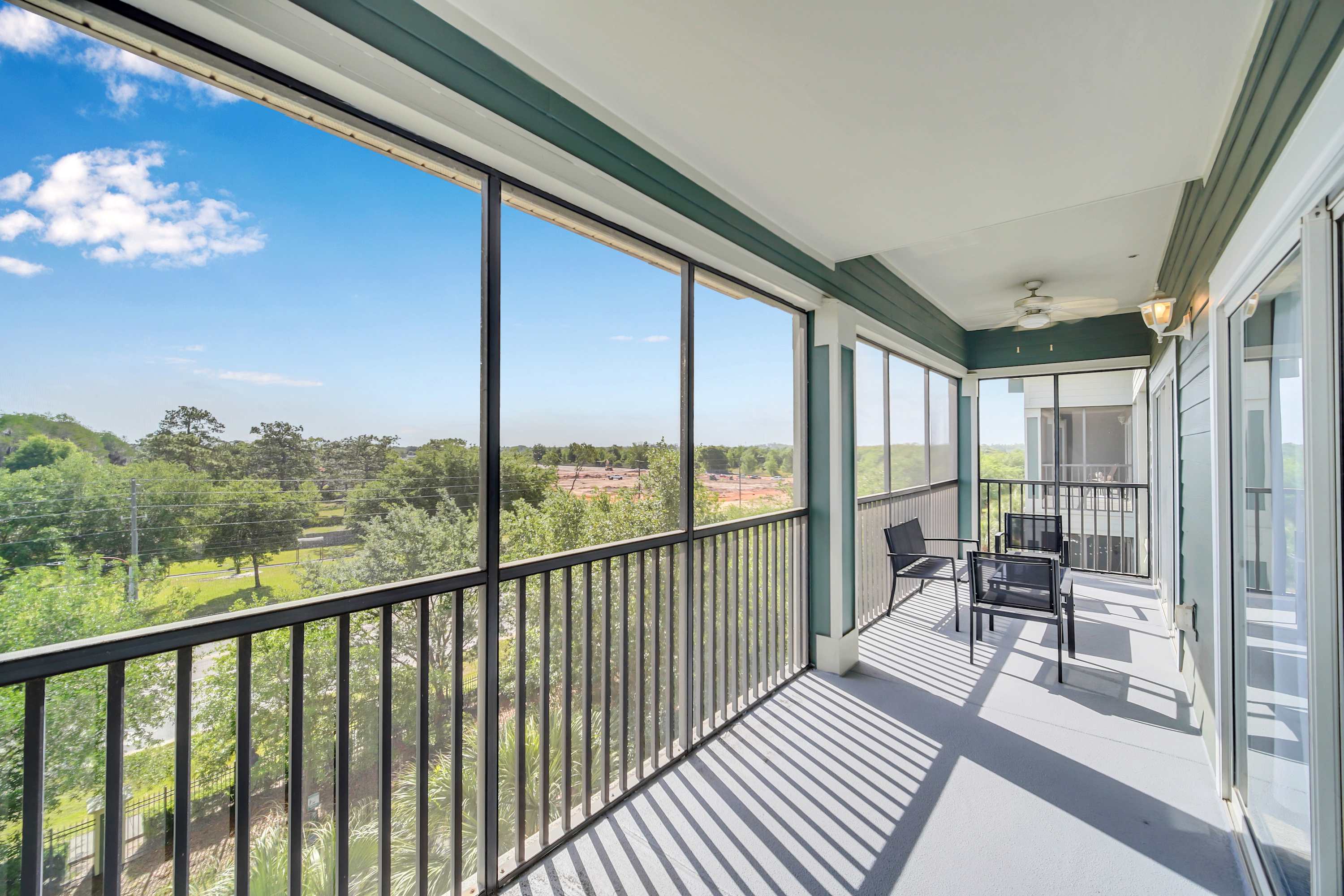 A spacious screened-in balcony with gray flooring and black railings, offering panoramic views of lush green trees and a distant building under a clear blue sky. Two black outdoor chairs and a small table are visible.