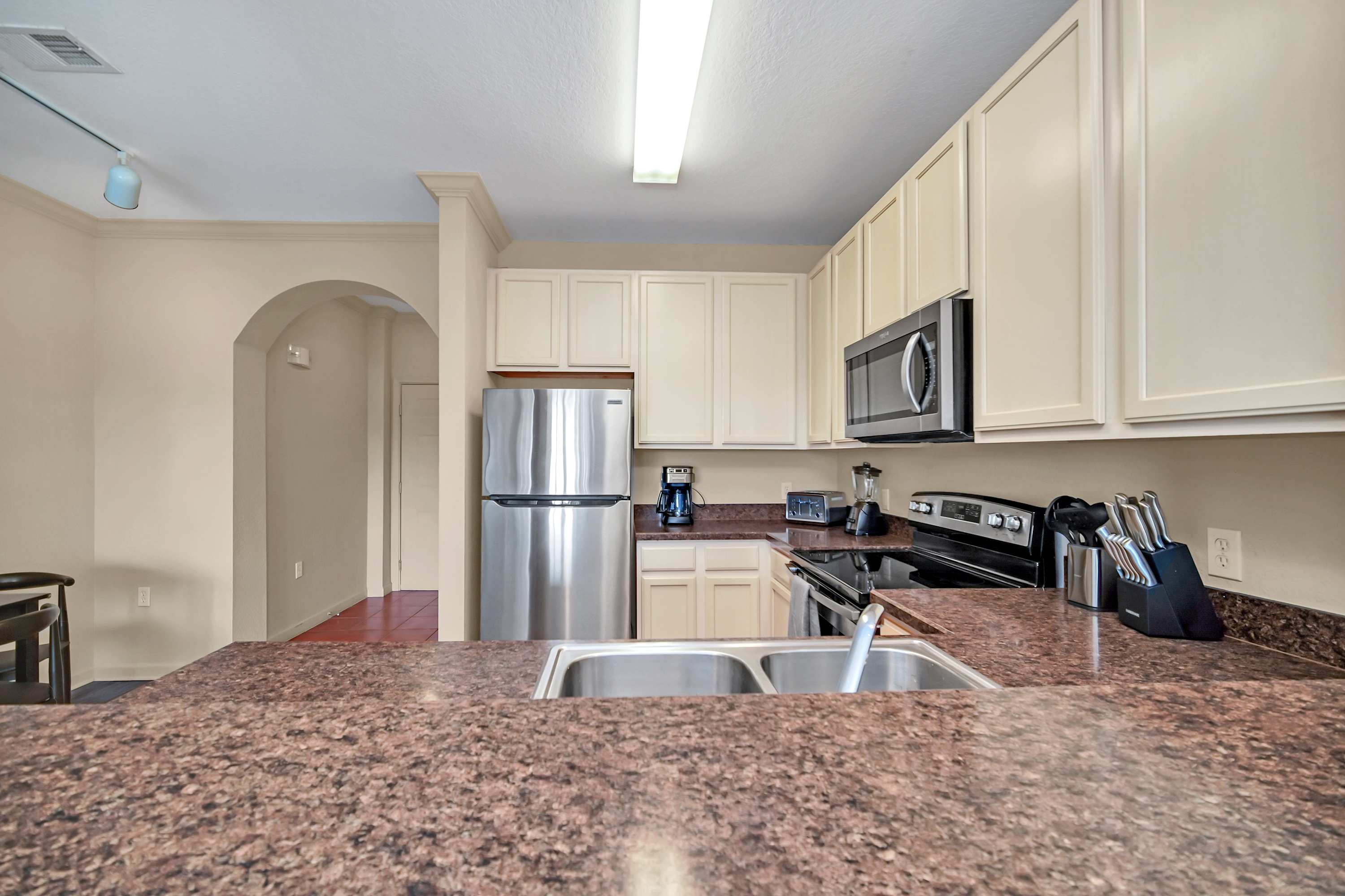 A close-up of the kitchen counter highlights a double stainless steel sink set into a speckled brown granite countertop. Various small appliances like a coffee maker, toaster, and blender are visible, with light cabinets above.