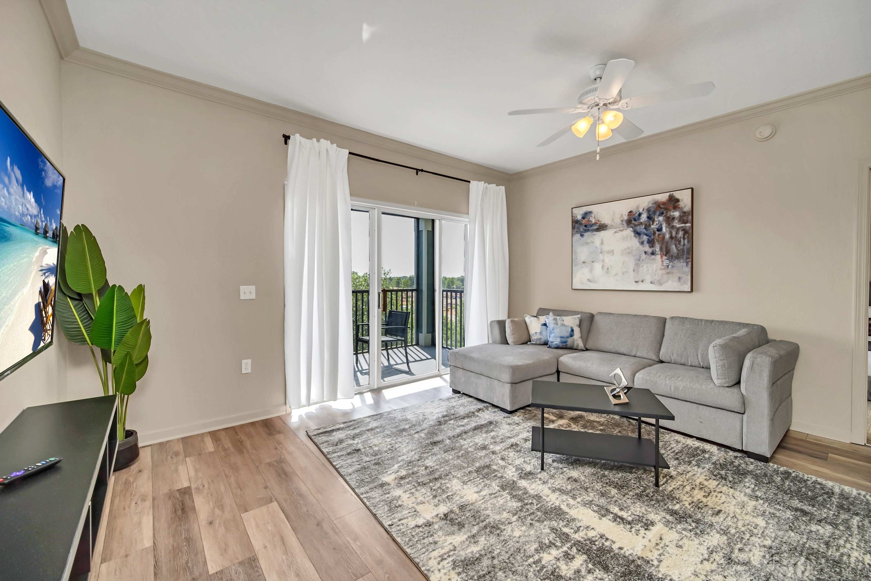 A bright living room features a light gray sectional sofa, a dark coffee table, and a textured area rug over wood flooring. A wall-mounted TV is visible, and a sliding glass door with white curtains opens to a balcony.