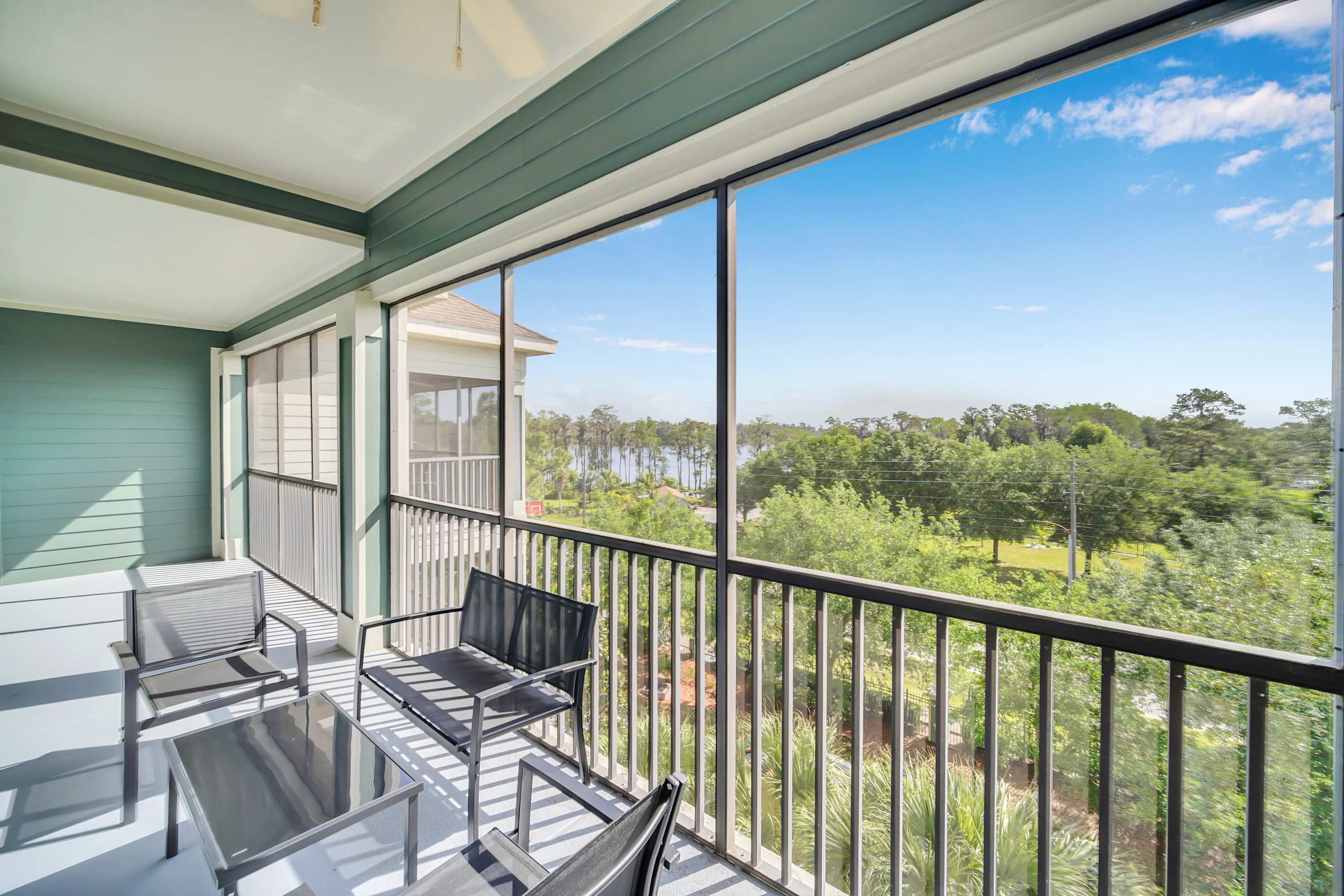 A screened balcony offers outdoor seating with a dark metal table and chairs, overlooking a lush landscape of trees and a distant body of water. The balcony features a dark railing and a light blue ceiling.