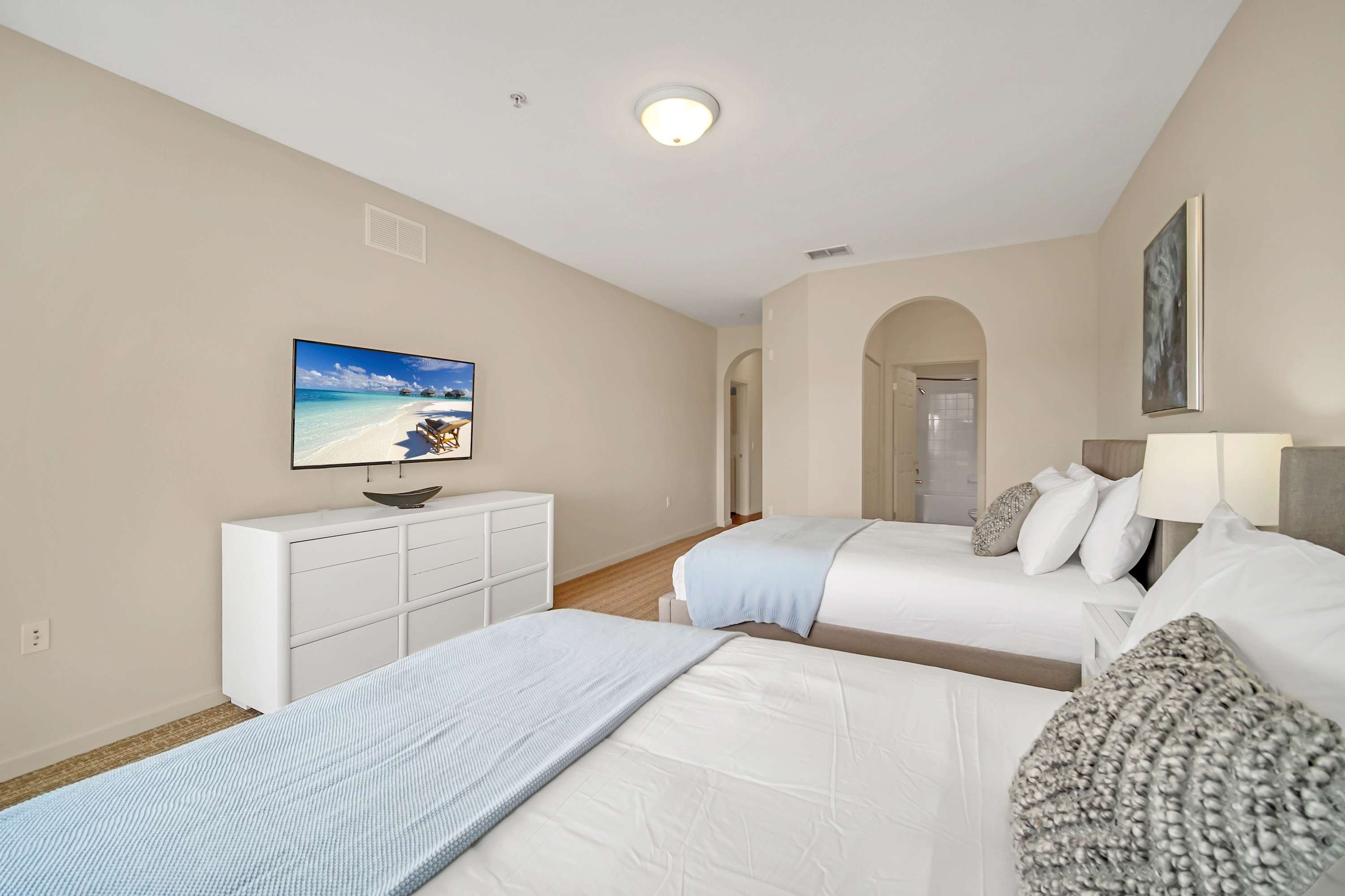 Another view of the bedroom with two queen beds, light-colored walls, and a wall-mounted flat-screen TV above a white dresser. An arched doorway in the background leads to an en-suite bathroom.
