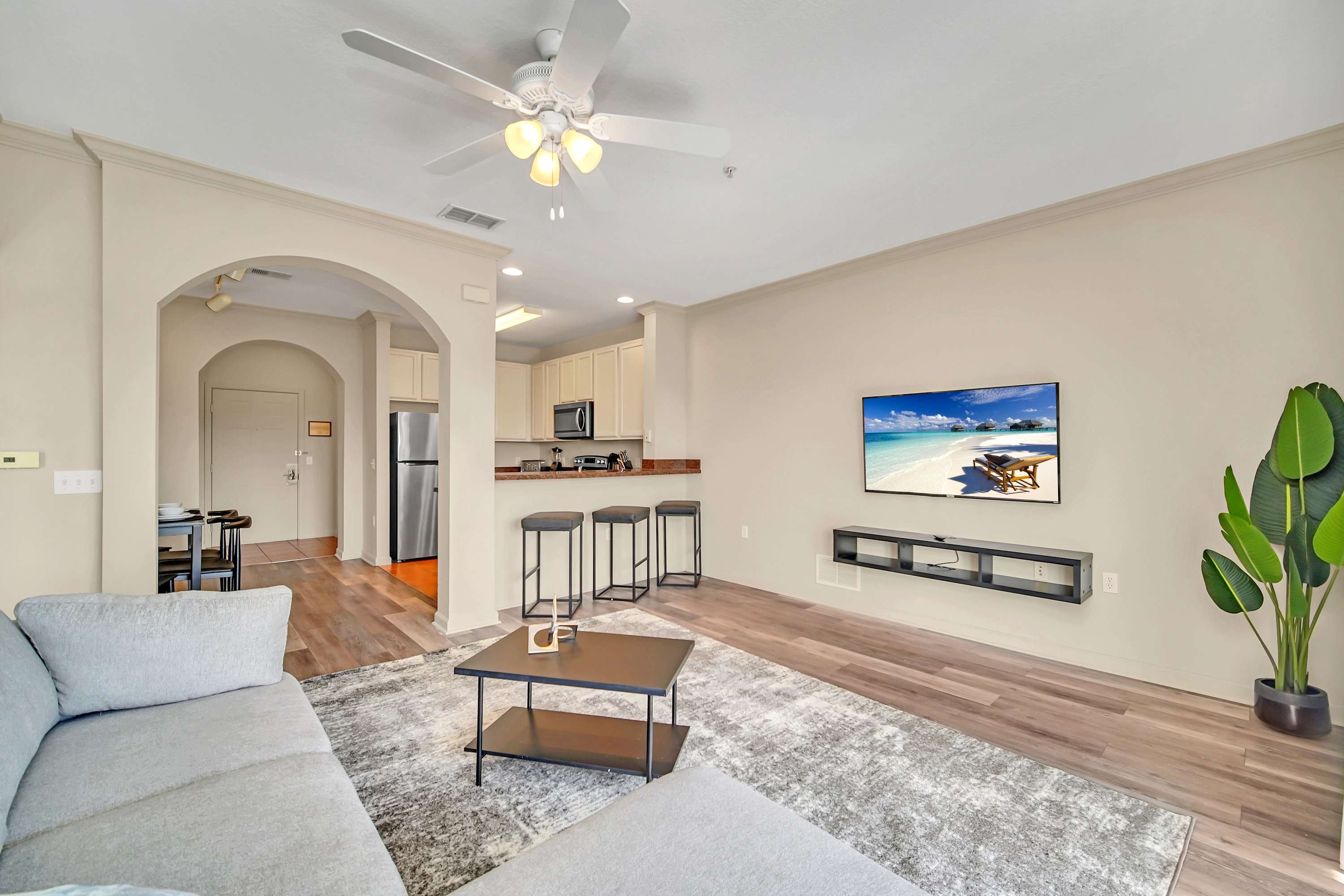 An open-concept living space features a light gray sectional sofa, a dark coffee table, and a large area rug. A wall-mounted TV is visible, and a breakfast bar with three stools separates the living area from the kitchen.