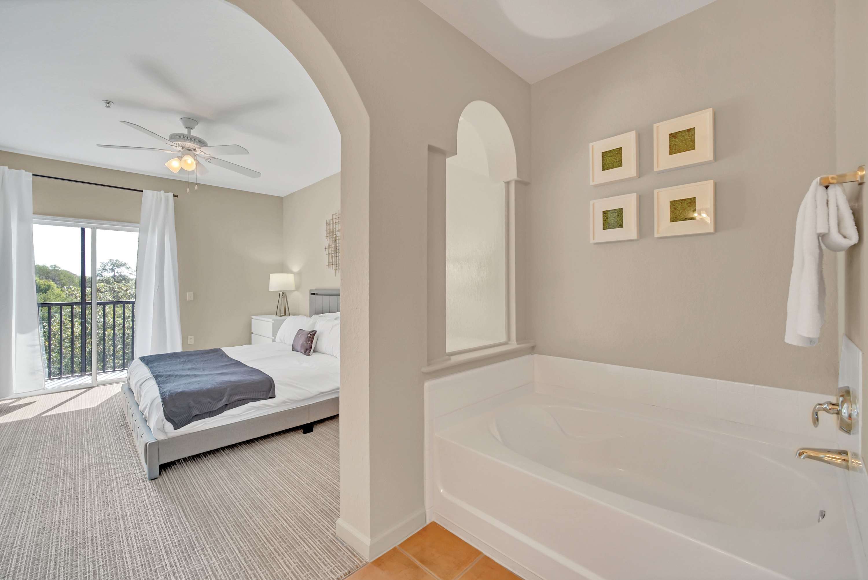 A modern bedroom featuring a queen-sized bed with white and gray bedding, a ceiling fan, and access to a private balcony with outdoor views. An arched doorway leads to a bathroom with a large white soaking tub and light gray walls.