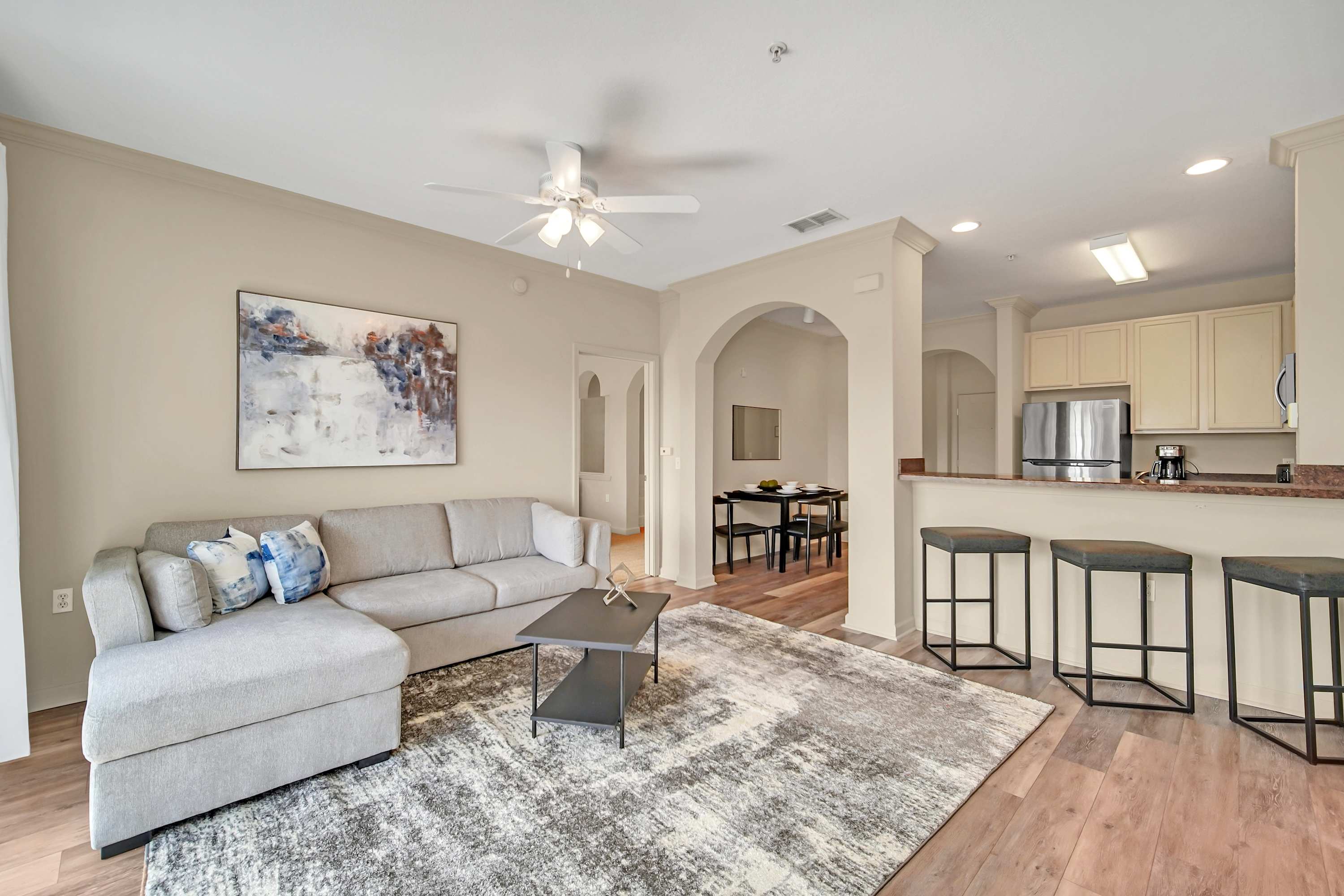A spacious living room features a light gray sectional sofa, a dark coffee table, and a large area rug, with an abstract painting on the wall. A breakfast bar with three stools connects to the kitchen, and an archway leads to the dining area.