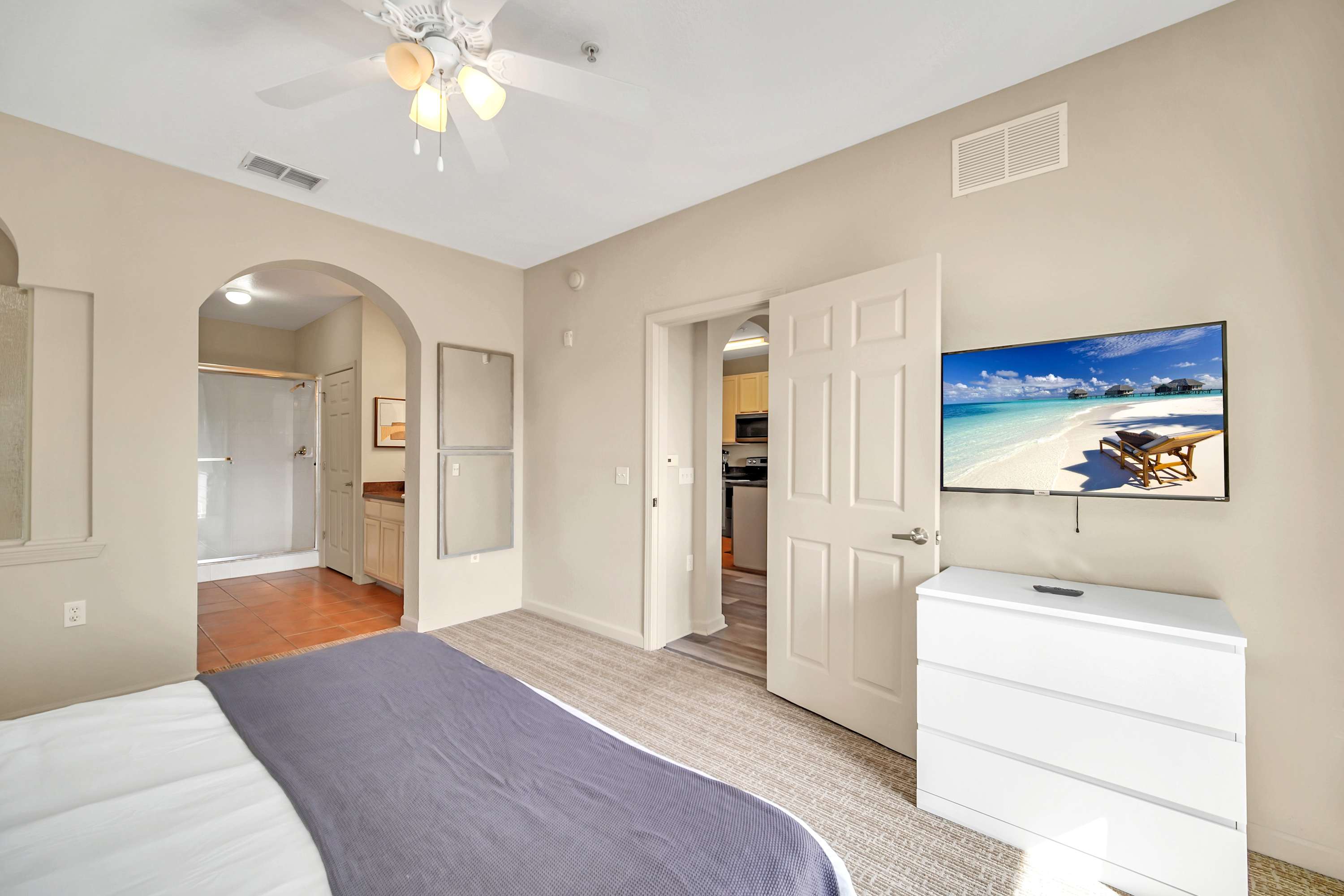 A cozy bedroom with a queen bed, light gray carpet, and a wall-mounted flat-screen TV above a white dresser. An arched opening leads to a bathroom, and a white door opens to a hallway or kitchen area.