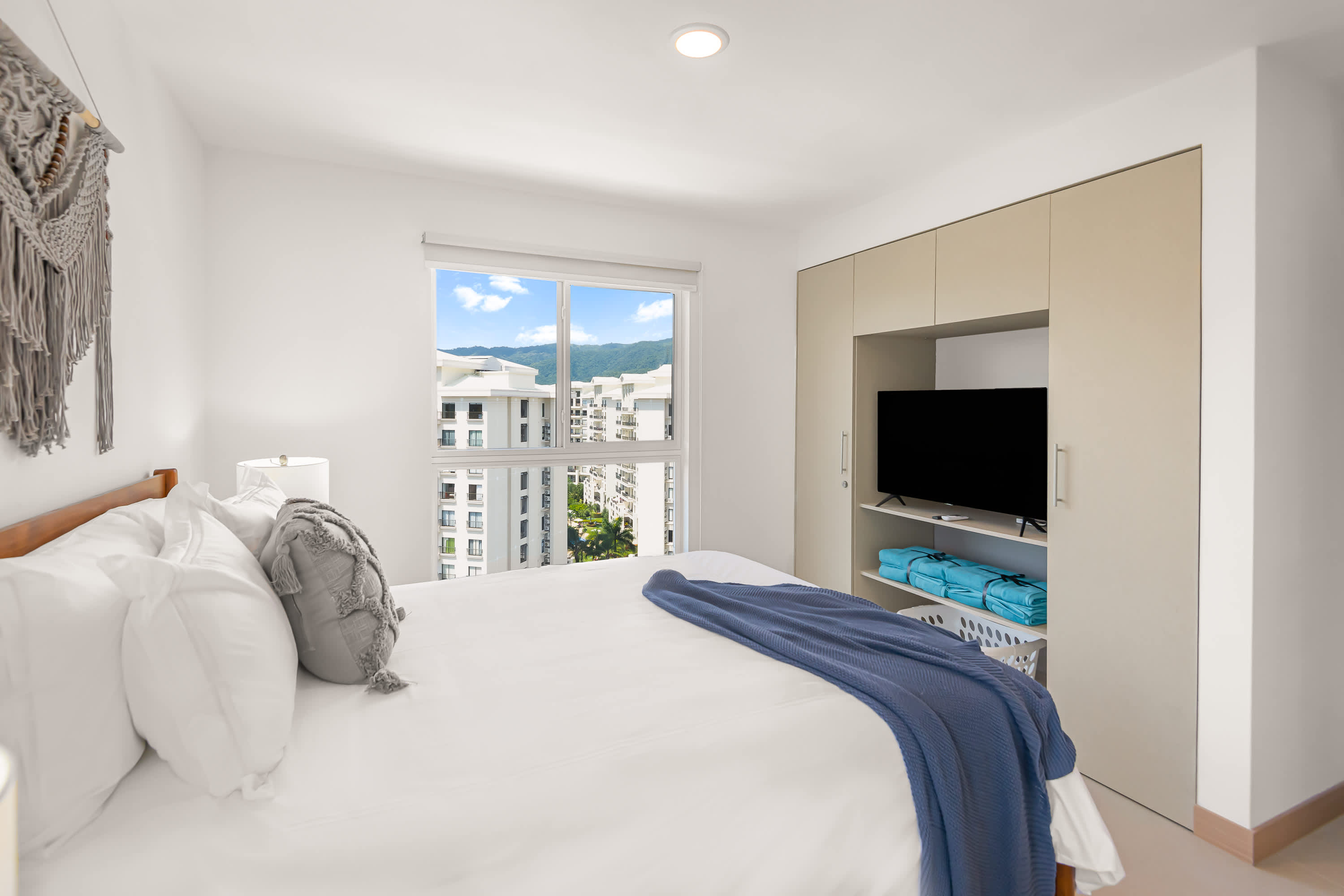 View of bedroom-1 showcasing a queen bed with white linens and a grey throw, facing a built-in closet with sliding doors and an integrated television. A large window offers views of surrounding buildings and mountains.