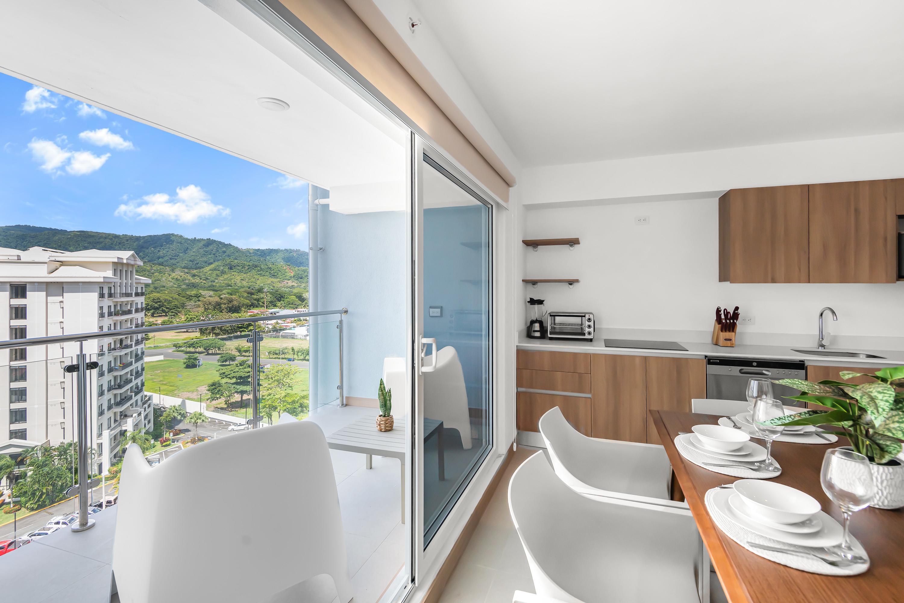 Modern open-concept living space featuring a dining table with white chairs, a compact kitchen with wood cabinetry, and a balcony offering expansive mountain and city views. The balcony has outdoor seating and a small plant.