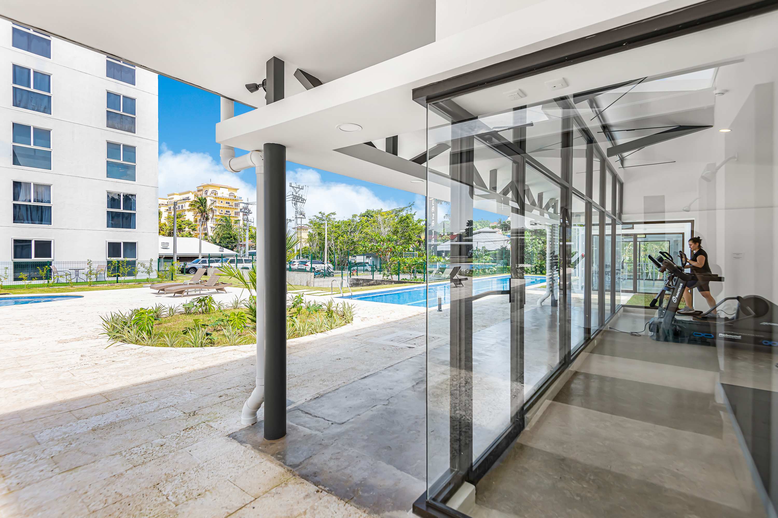 A bright gym with large glass walls offers views of the outdoor pool and patio area, featuring a treadmill and other exercise equipment. The modern building has white walls and a sleek design.