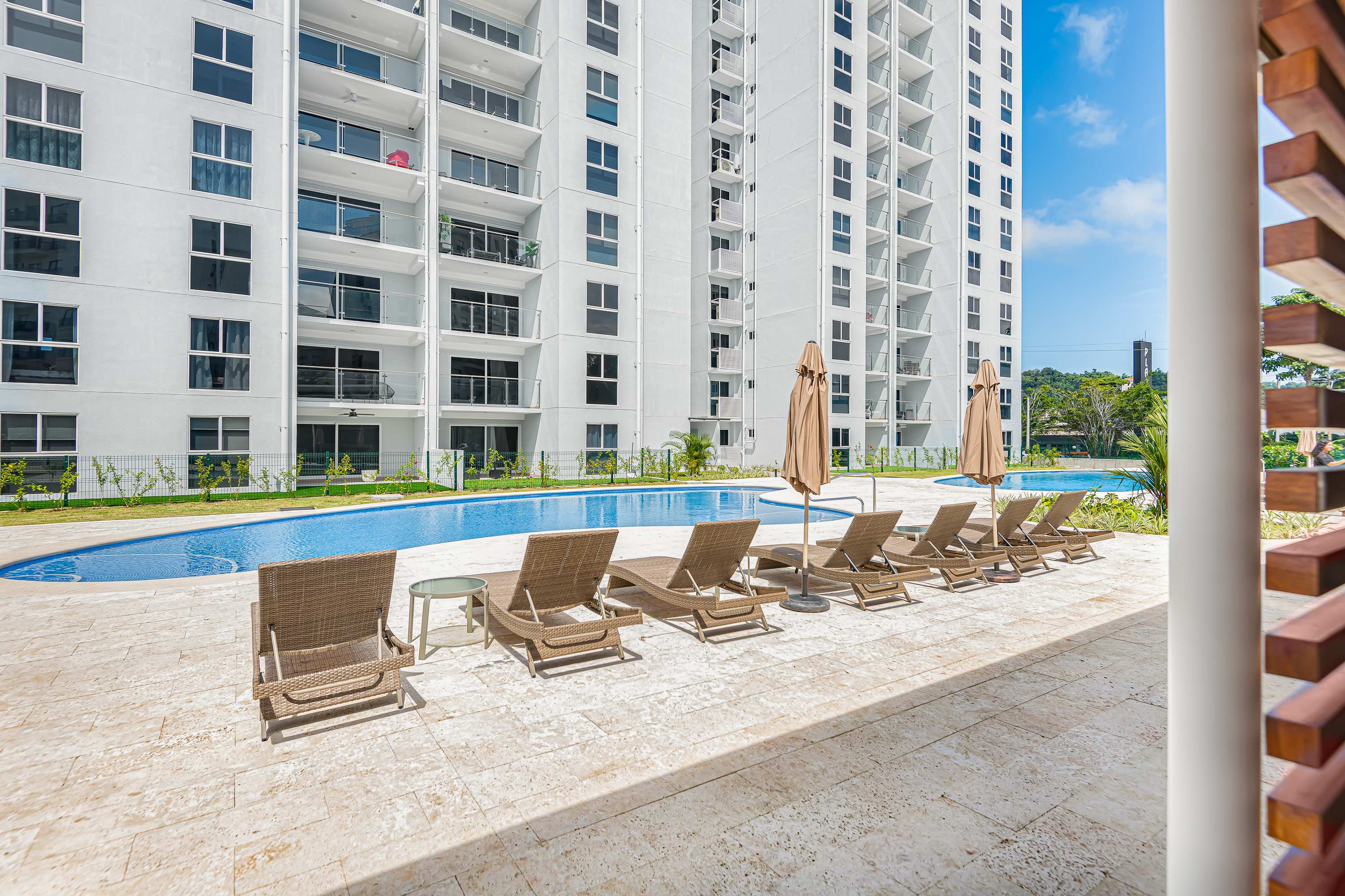 A large, irregularly shaped communal swimming pool is surrounded by numerous brown wicker lounge chairs with umbrellas on a light-colored paved deck. The modern white condominium building rises in the background.