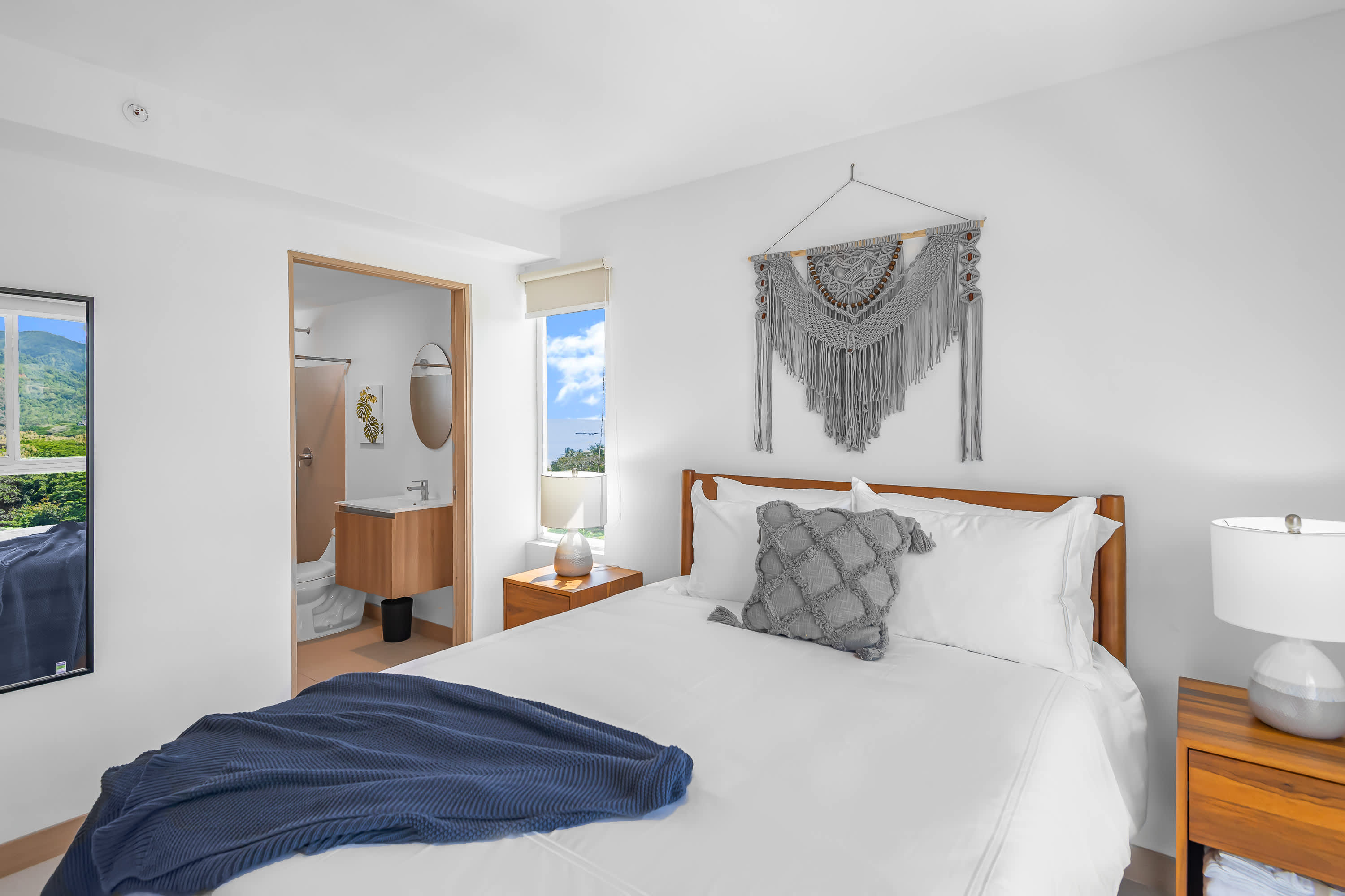 Serene bedroom-1 with a queen-sized bed, white bedding, and a grey textured throw pillow, featuring a macrame wall hanging above the wooden headboard. An open doorway leads to an ensuite bathroom with a modern vanity.