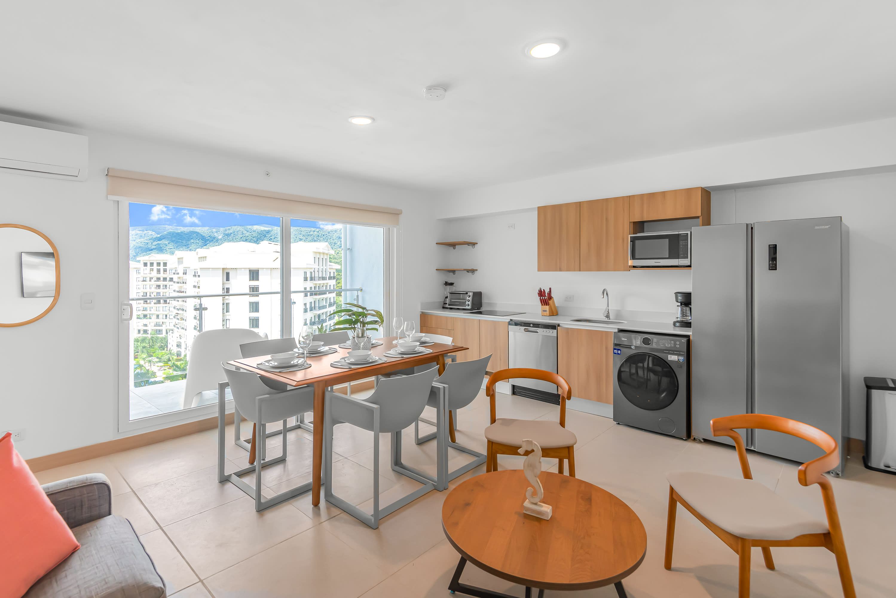Open-plan living space featuring a gray sofa, a dining table set for six, and a modern kitchen with light wood cabinets, all bathed in natural light from the large balcony doors.