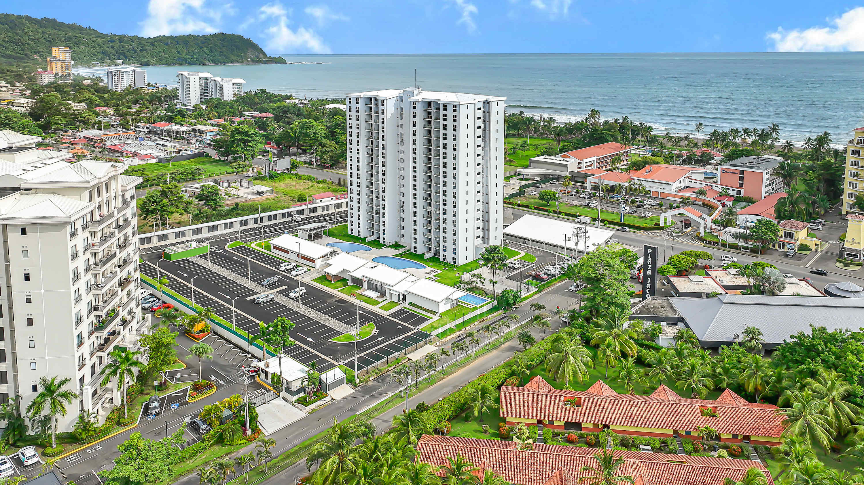 Aerial view of a tall, modern condominium building with multiple balconies, a large parking lot, and a swimming pool, situated near the ocean and surrounded by other buildings and lush landscape.