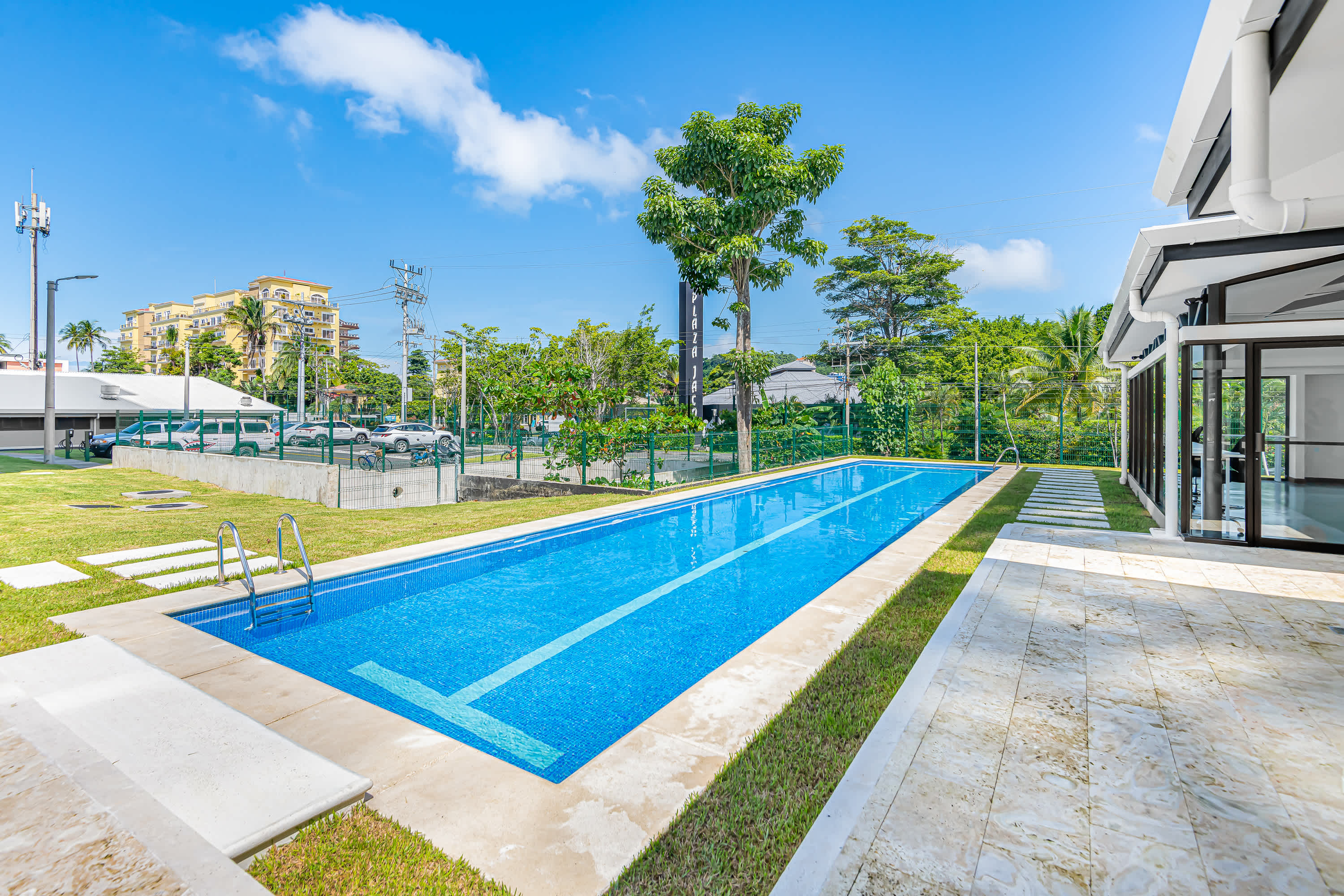 A long, narrow rectangular swimming pool with blue tiled interior is set in a grassy area, surrounded by light-colored paving stones and tropical trees under a bright blue sky. Steps with handrails lead into the pool.