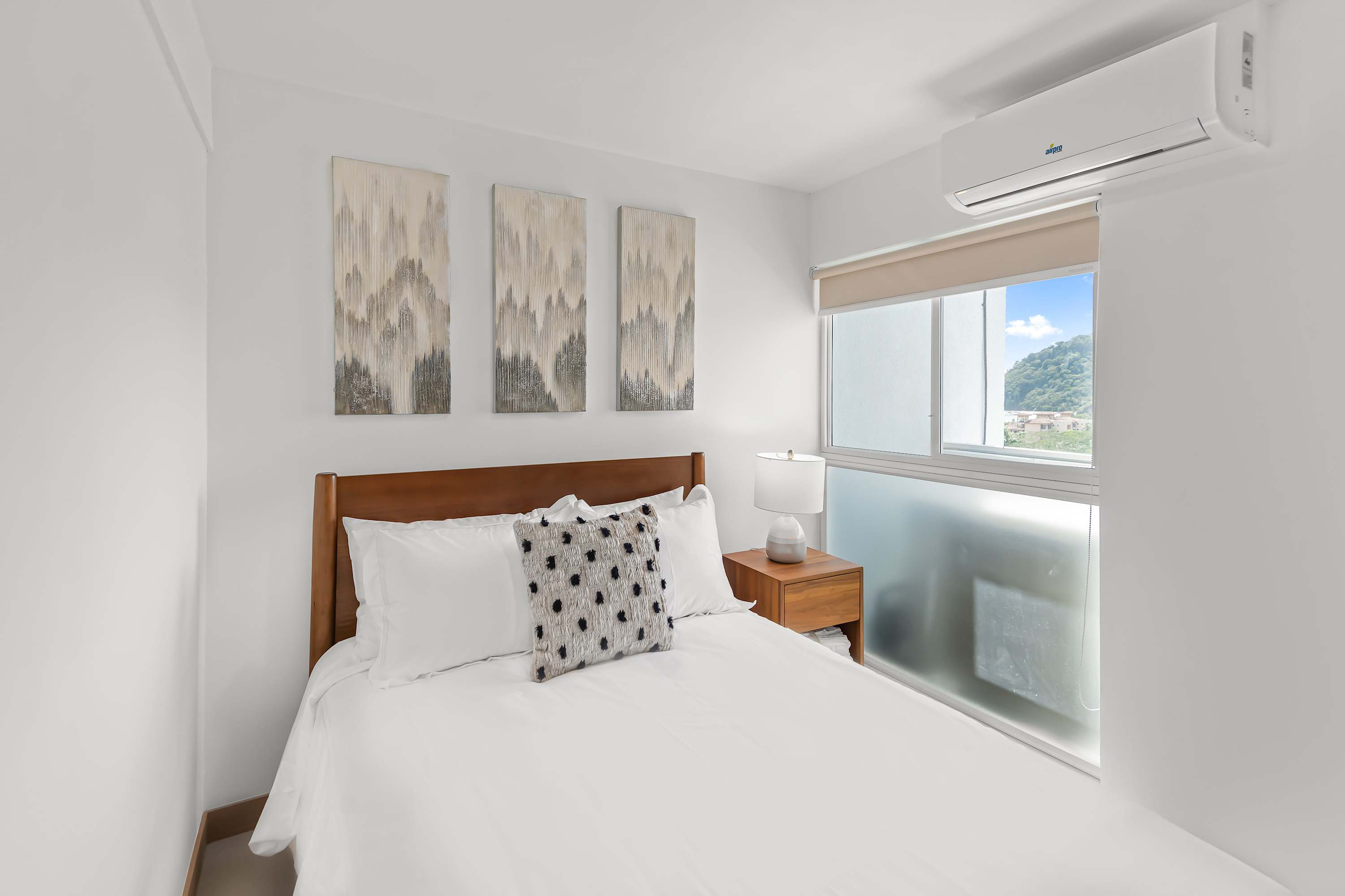 Another perspective of bedroom-3, highlighting the full-sized bed with white linens and a patterned pillow, and the three abstract art pieces on the wall. A window offers a glimpse of the outside view, and an air conditioning unit is mounted above.