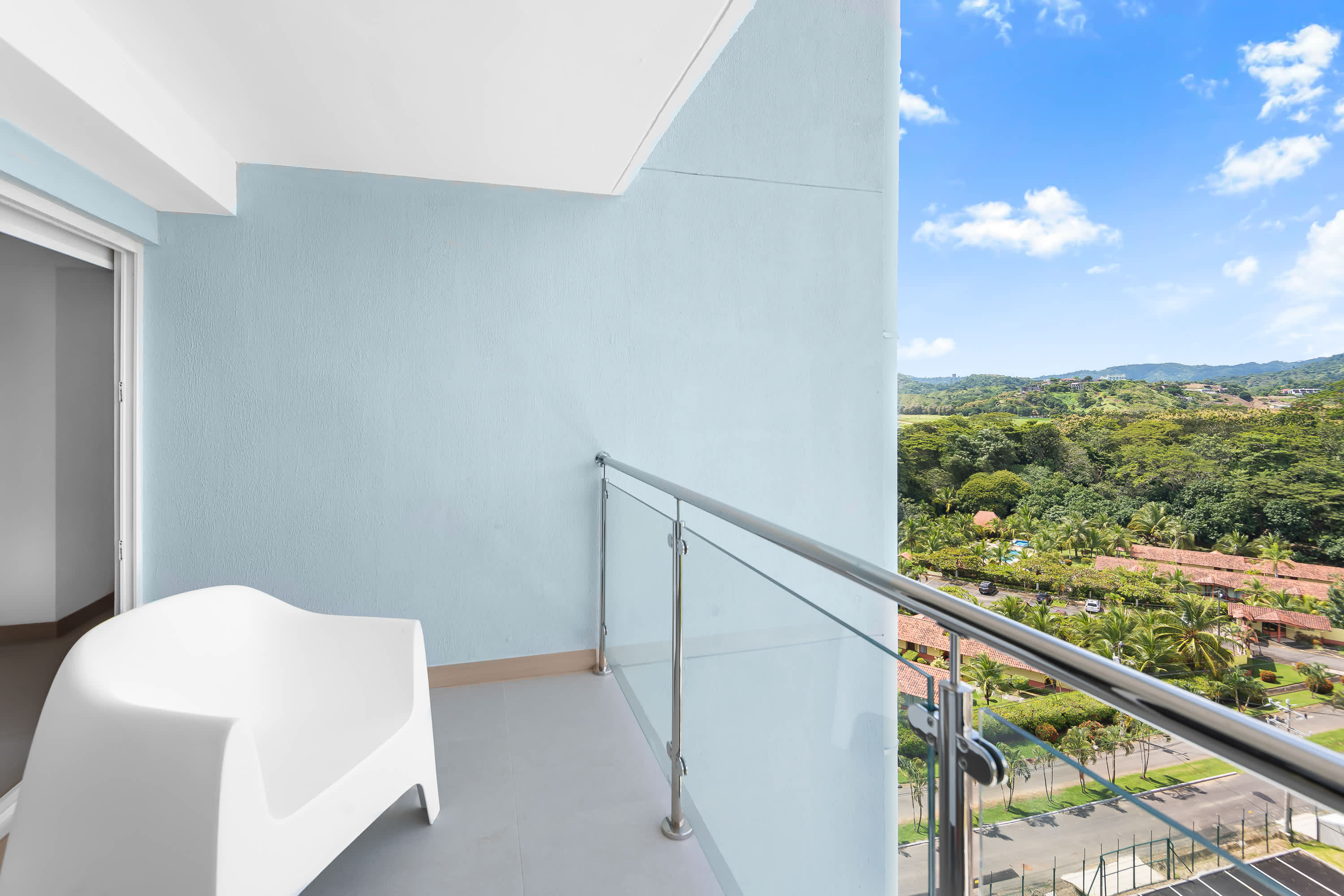 Private balcony with a glass railing and a white chair, offering a panoramic view of lush green mountains and distant buildings under a bright blue sky.