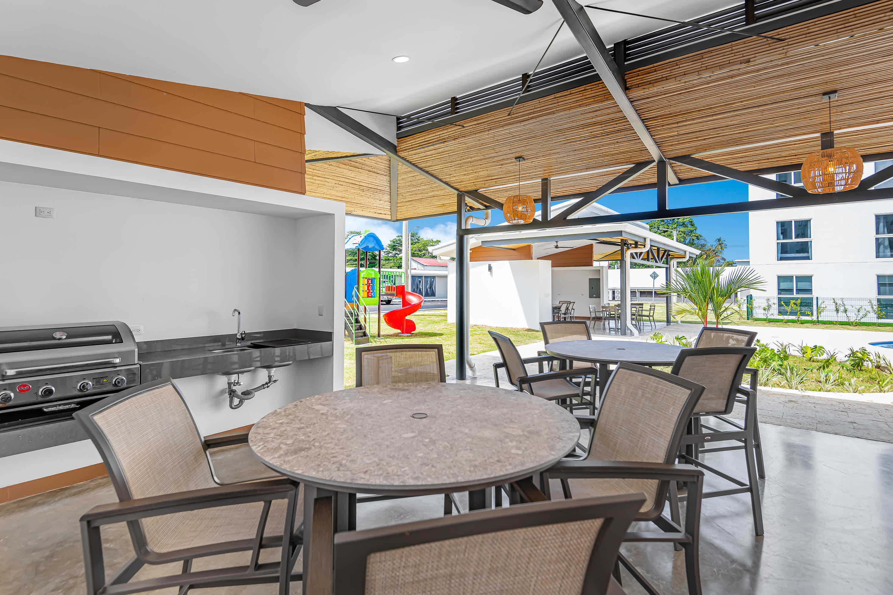 An outdoor covered dining area features a built-in grill, sink, and round tables with chairs, all under a slatted wooden ceiling. A colorful children's playground is visible in the background.