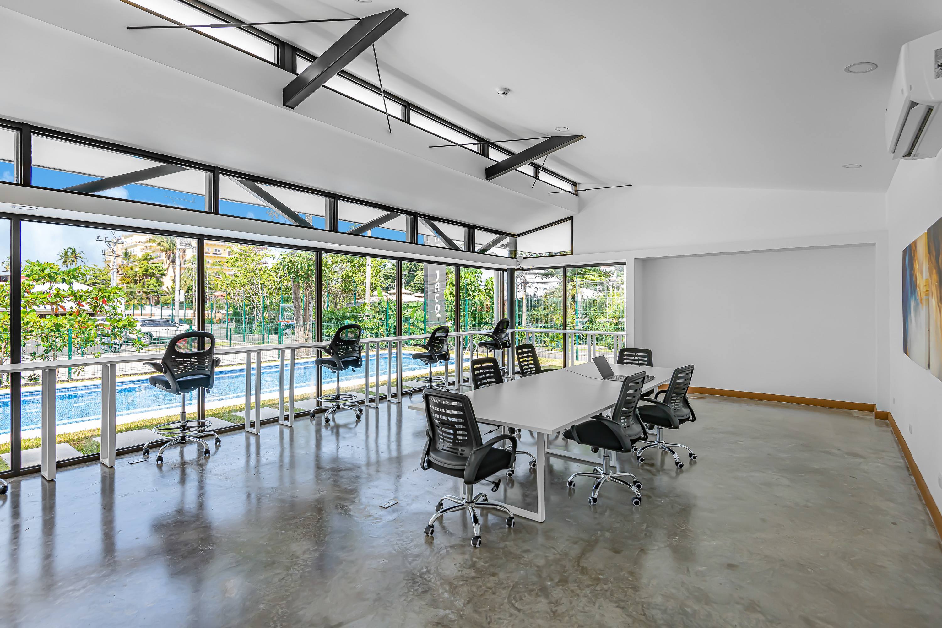 A modern co-working space features a long white conference table with black office chairs, and individual workstations along a window overlooking a pool. The room has a high ceiling with exposed black beams and polished concrete floors.