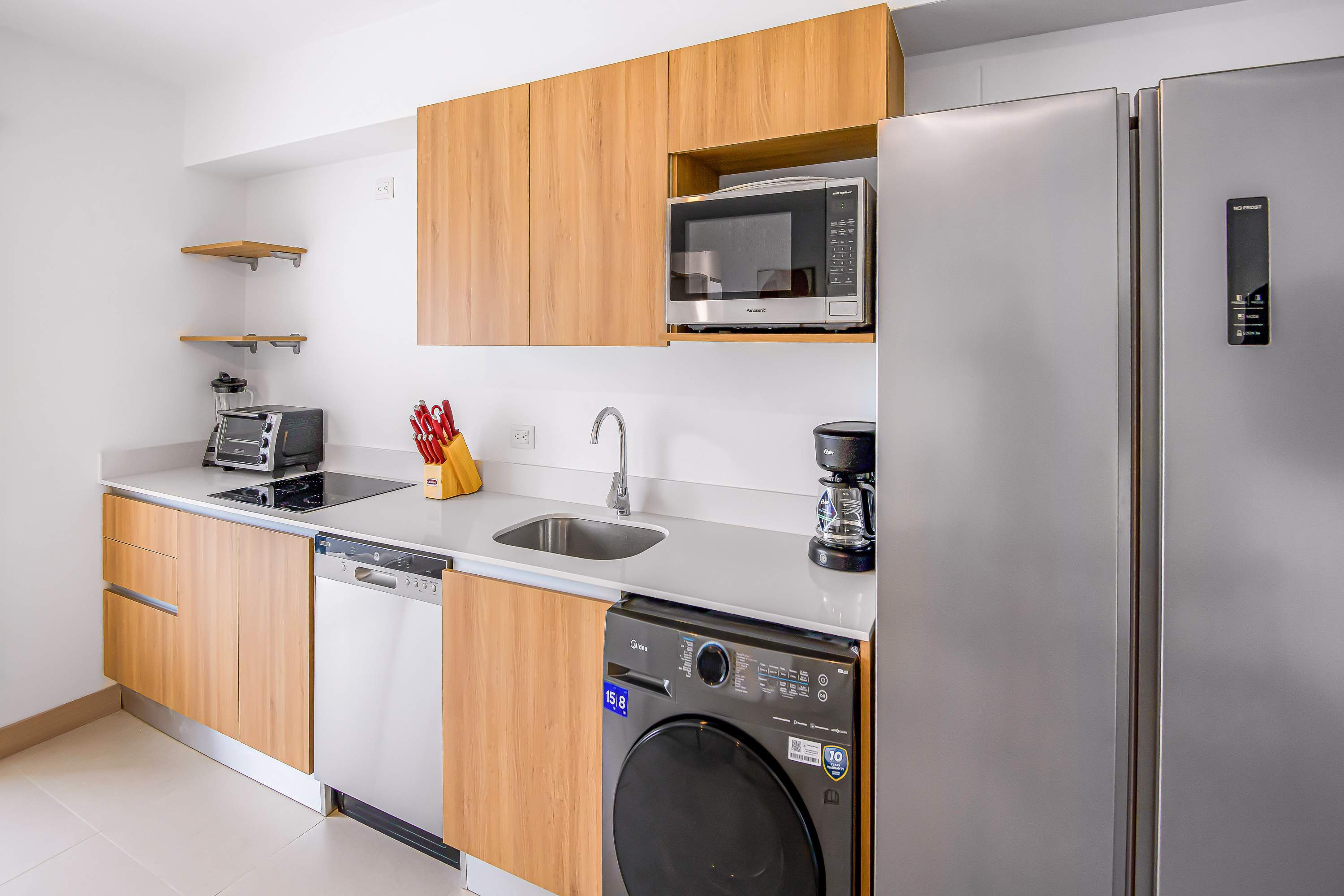 Modern kitchen area showcasing light wood cabinets, white countertops, and stainless steel appliances, including a large refrigerator and a built-in microwave, with a view towards the living area.
