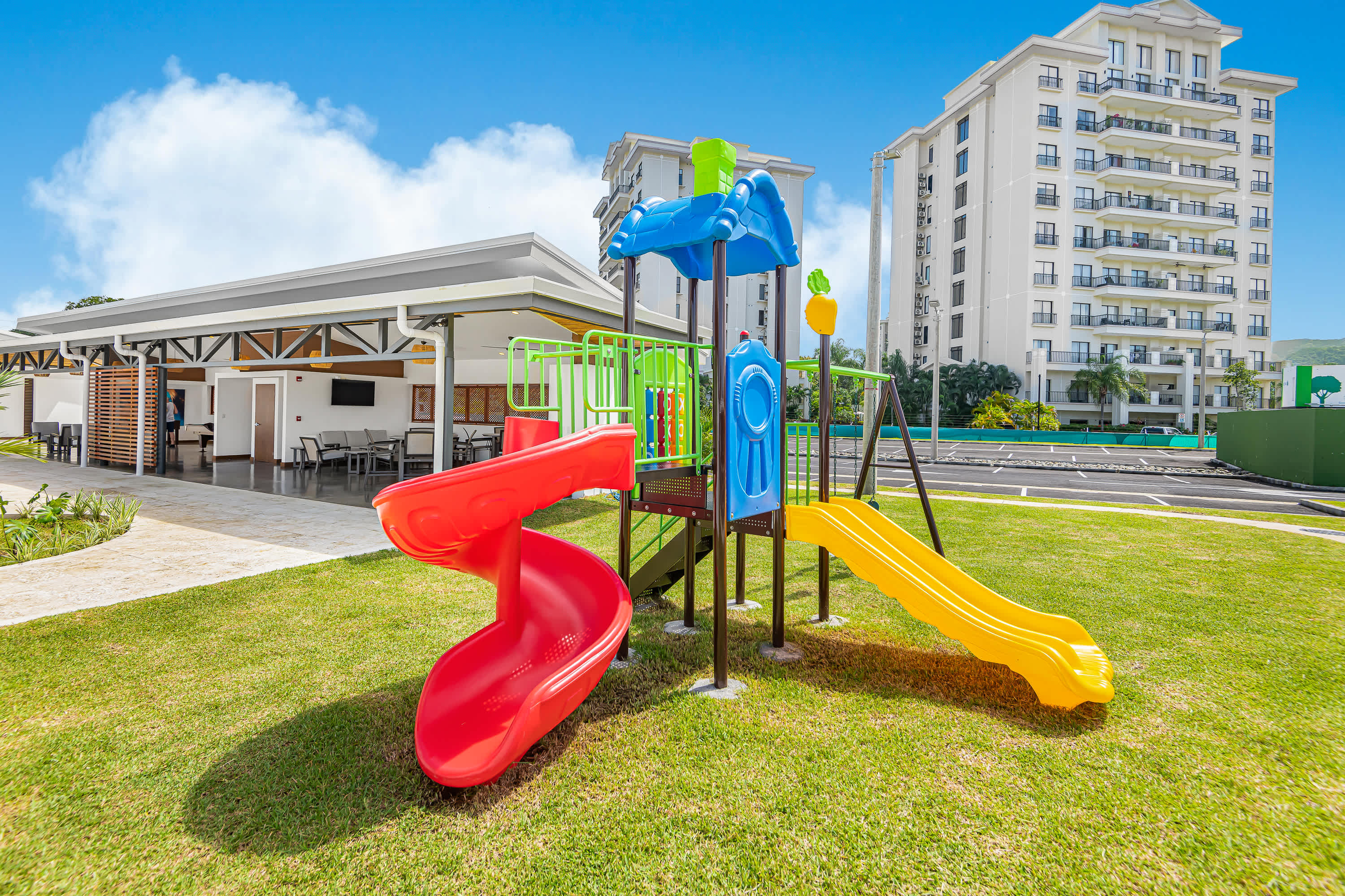 A vibrant children's playground with red and yellow slides, green and blue climbing structures, and swings is set on a lush green lawn. A covered outdoor area and the condominium building are visible in the background.