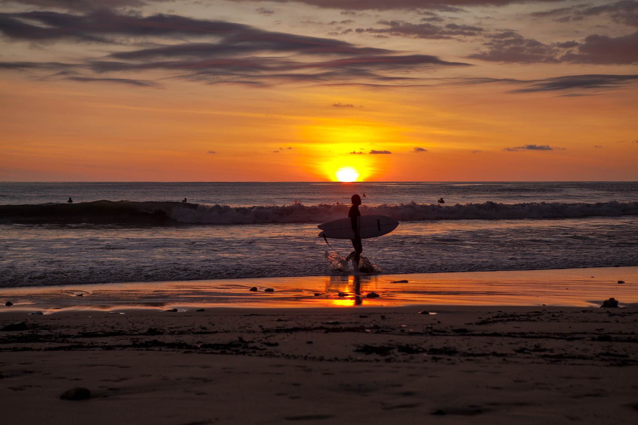 A lone surfer walks along a sandy beach at sunset, carrying a surfboard, with the vibrant orange sun dipping below the horizon over the ocean. Waves crash gently, and other surfers are visible in the distance.