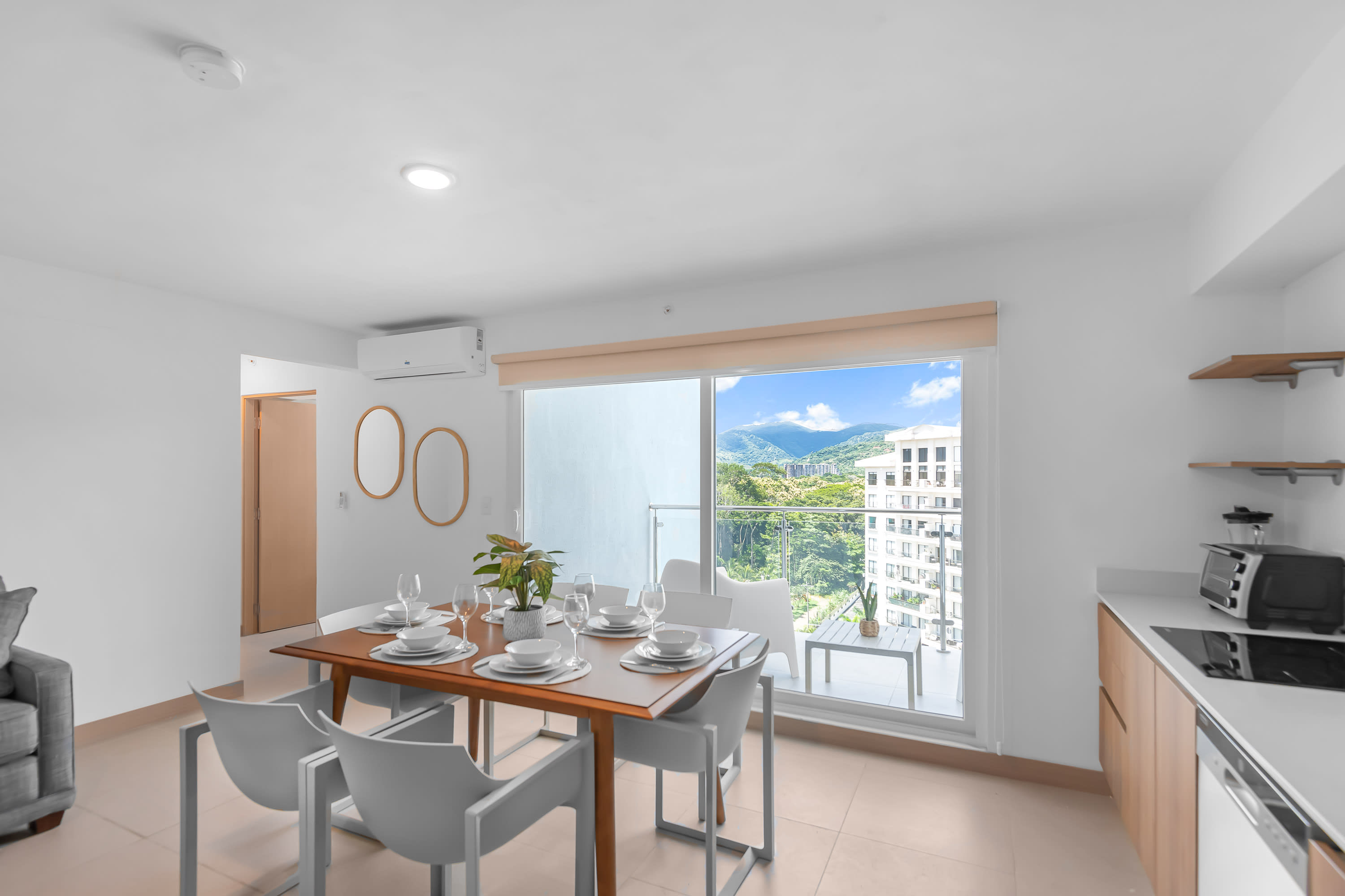 Bright open-plan dining area with a wooden table set for six, white chairs, and a view of the balcony and mountains through large windows. A modern kitchen with wood cabinets and white countertops is visible to the right.