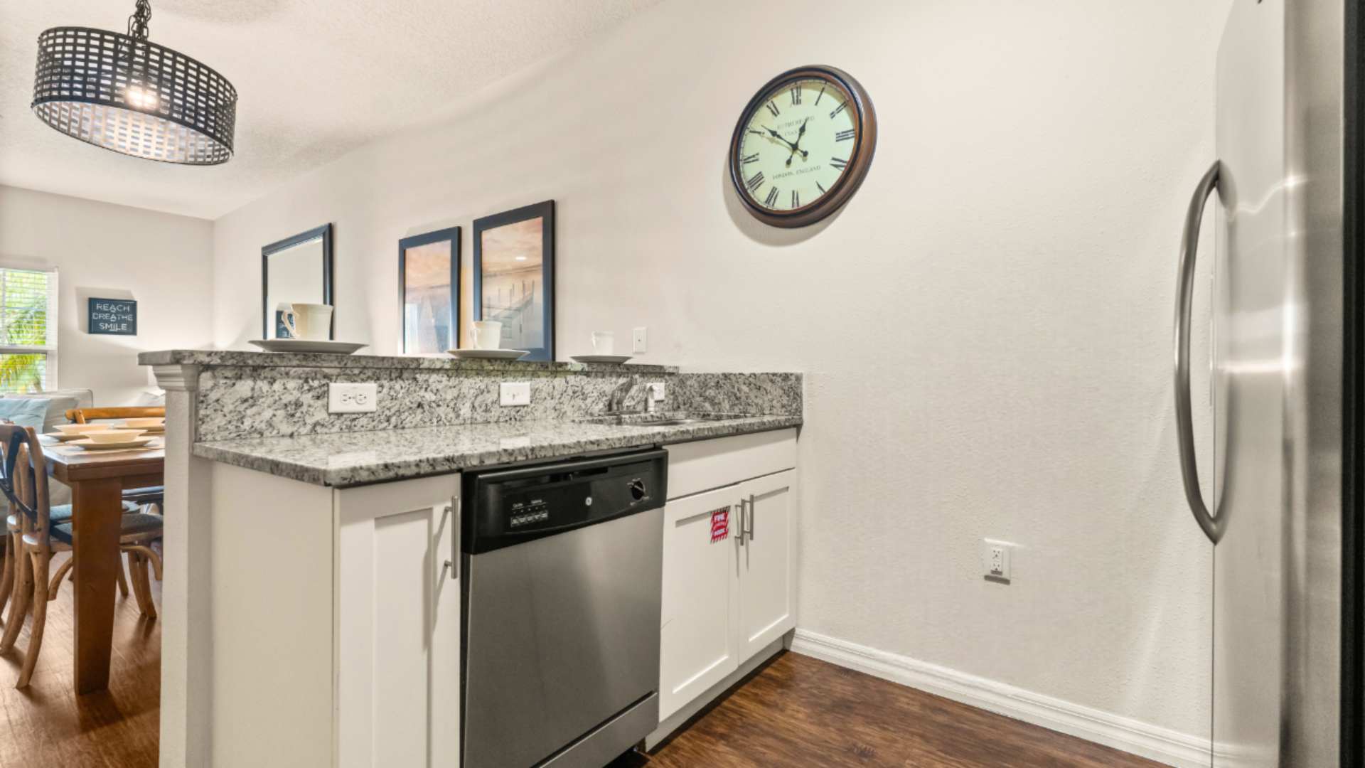 Kitchen bar area with granite countertops, a stainless steel dishwasher, and a double sink, illuminated by a modern black pendant light. A large round wall clock hangs on the white wall.