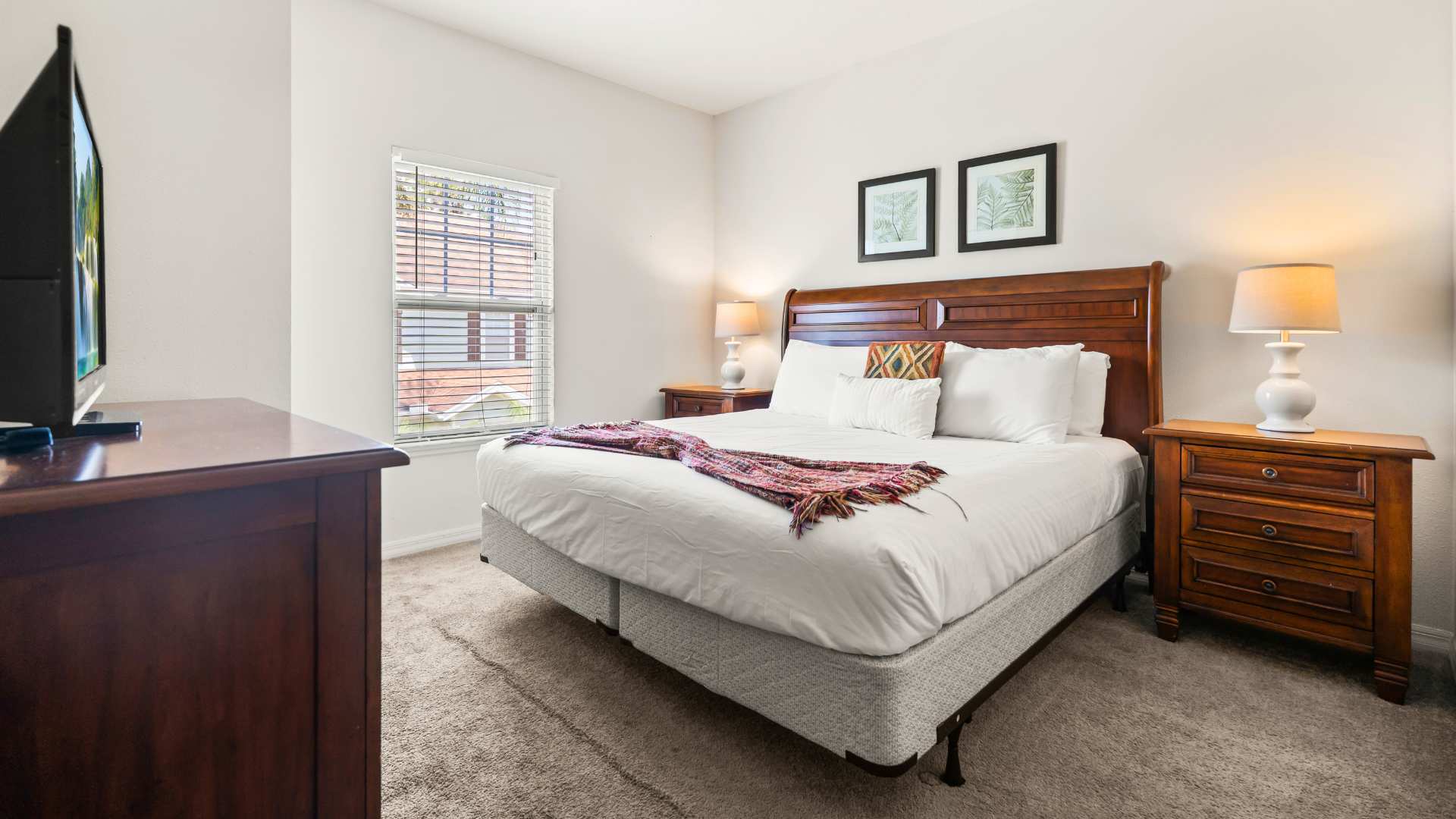 Bedroom with a queen-sized bed, dark wooden headboard, and matching nightstands with lamps. A dark wood dresser with a TV is positioned opposite the bed, and a window with blinds provides natural light.
