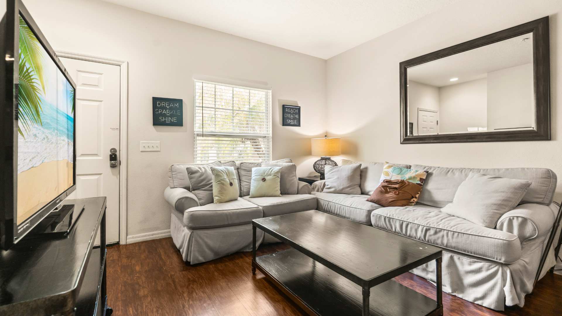 A bright living room features dark hardwood floors, a comfortable light gray sectional sofa adorned with decorative throw pillows, and a sleek black coffee table. A flat-screen TV rests on a dark console, positioned opposite a window with white blinds.