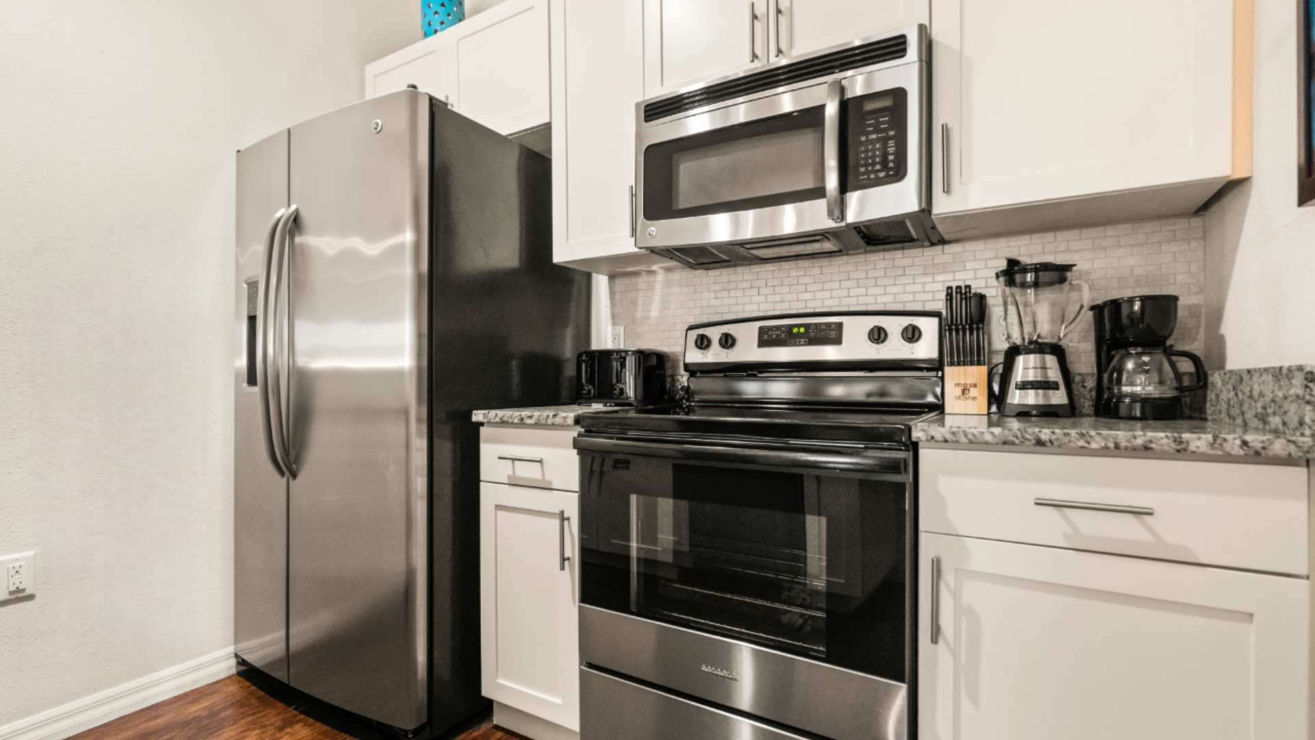 Full view of the modern kitchen featuring a stainless steel French door refrigerator, electric range, and microwave, complemented by white shaker cabinets and granite countertops. A toaster, coffee maker, and blender are on the counter.