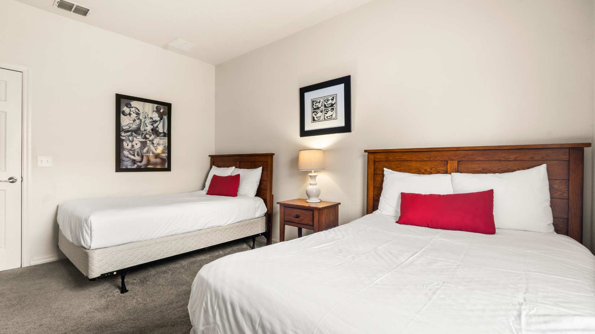 Bedroom with two twin beds, each with a wooden headboard and red accent pillow, separated by a nightstand with a lamp. Framed Mickey Mouse artwork hangs on the wall, and a white closet door is visible.