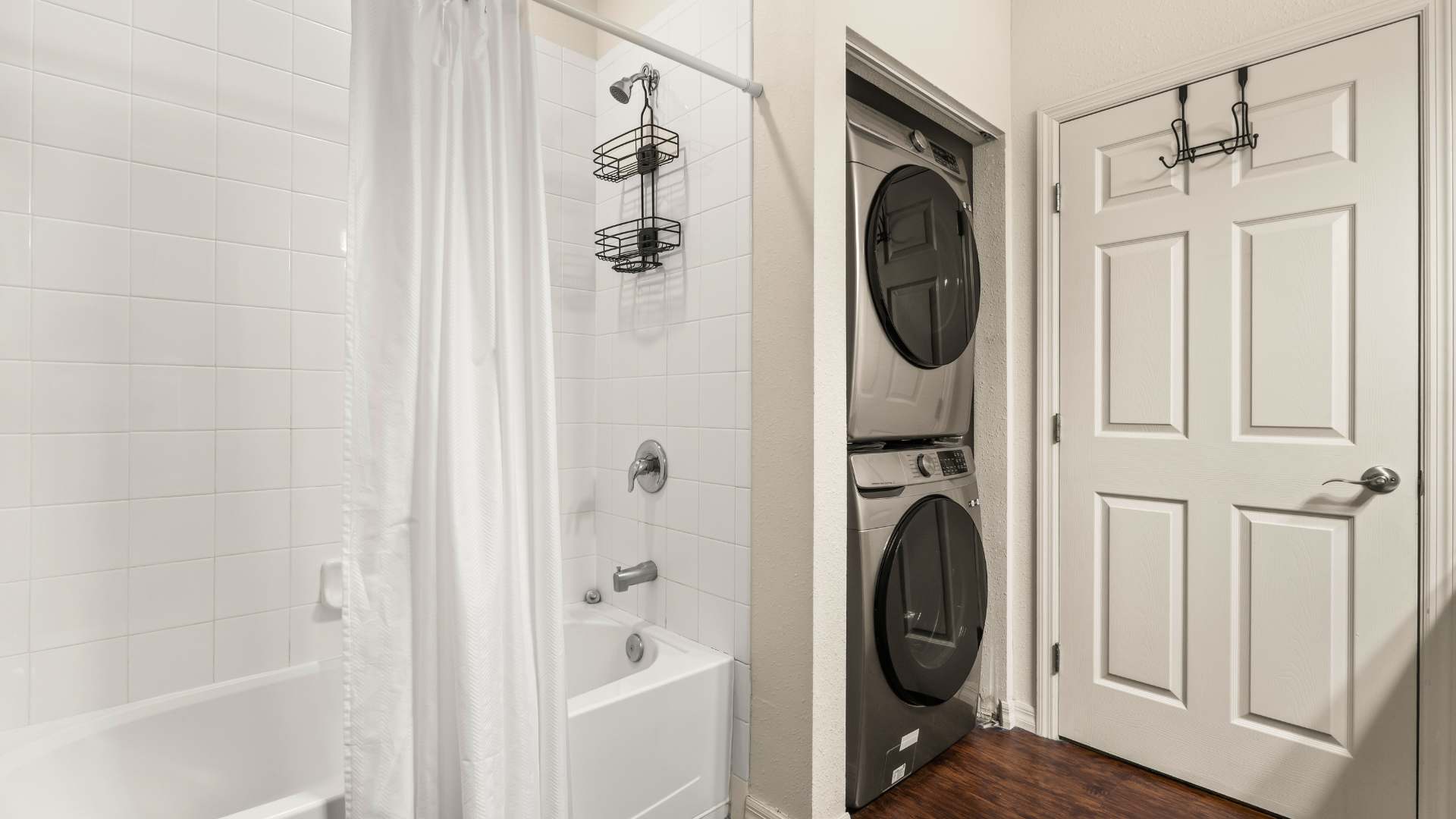 Clean bathroom featuring a white tiled shower-over-tub with a white curtain, and a stacked washer and dryer unit in a nearby closet, all set against dark wood flooring.