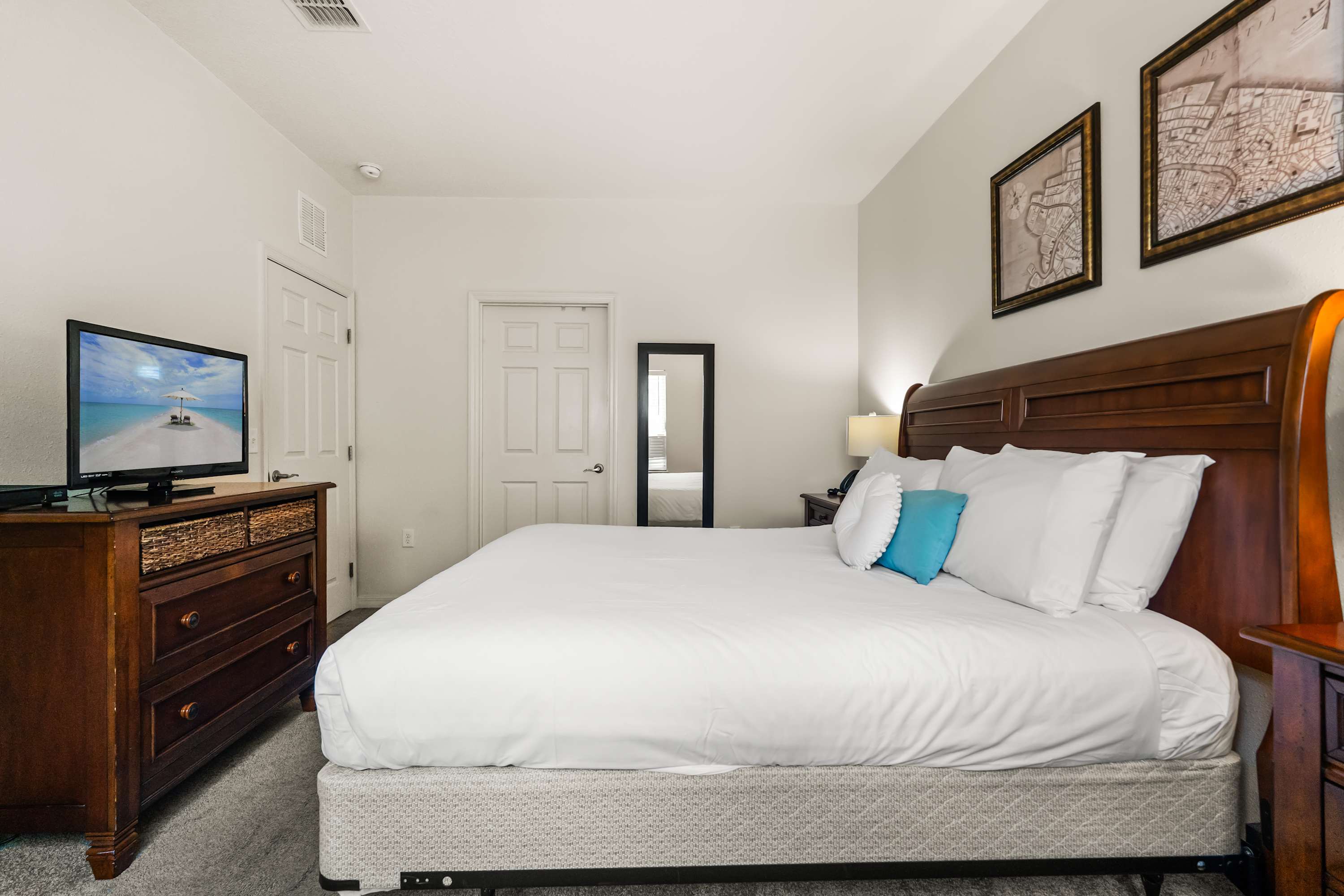 Spacious bedroom with a king-sized bed featuring a tall wooden headboard, white bedding with a blue accent pillow, and a dark wood dresser with a TV. Two framed maps adorn the wall above the bed.